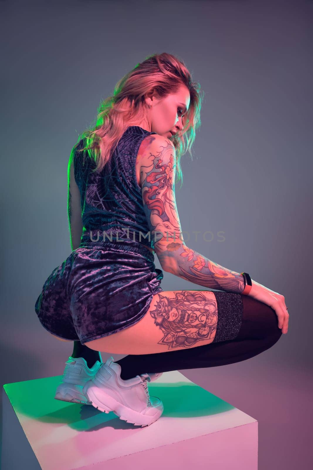 Beautiful young blonde tattoed girl in blue velour booty shorts and t-shirt, making squats on top of big cube, showing off her amazing booty