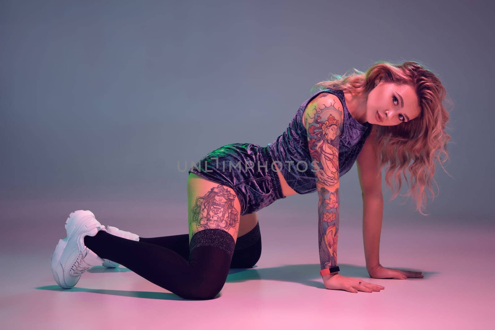 Young beautiful caucasian blonde girl with tattoo on her body, wearing velour blue booty shorts and black stockings dancing twerk on her knees, pink background