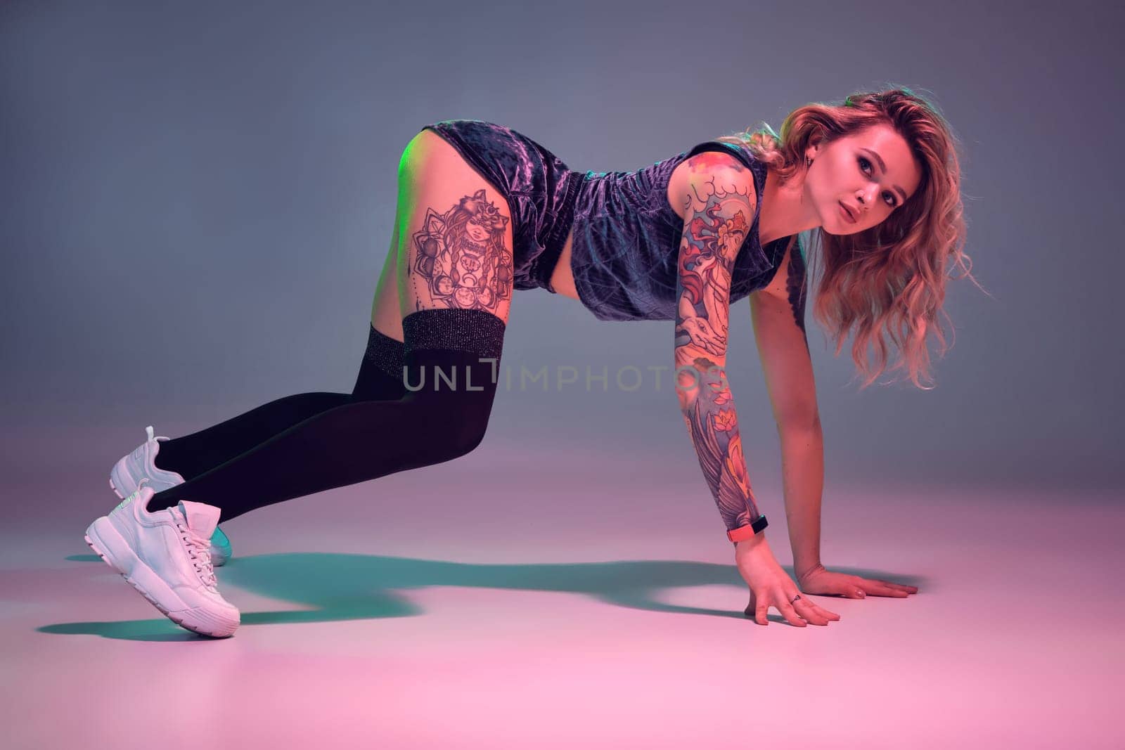 Young beautiful caucasian blonde girl with tattoo on her body, wearing velour blue booty shorts and black stockings dancing twerk, standing on four limbs, pink background