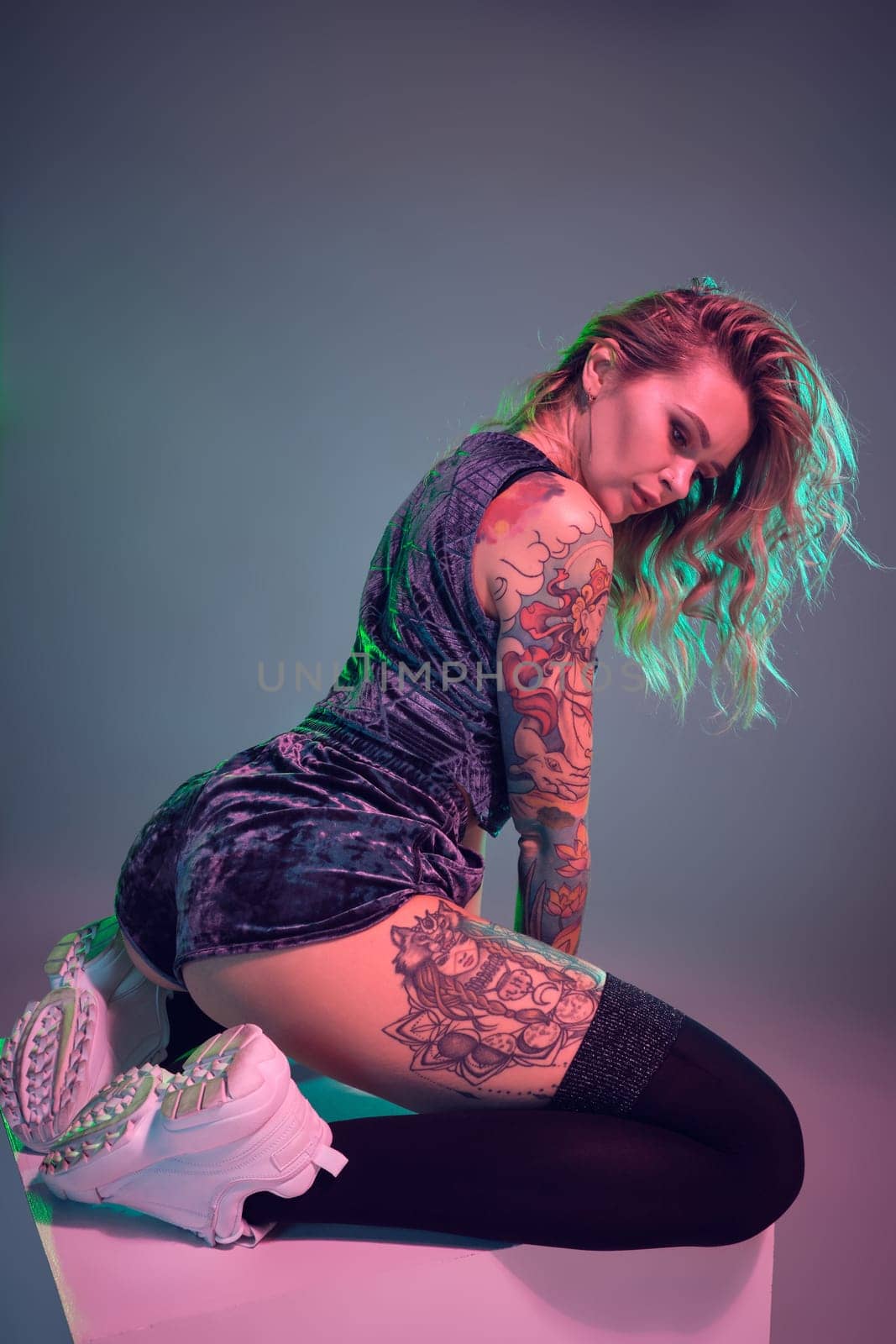 Beautiful young blonde tattoed girl in blue velour booty shorts and t-shirt, she sits on top of big cube, showing off her amazing booty from behind