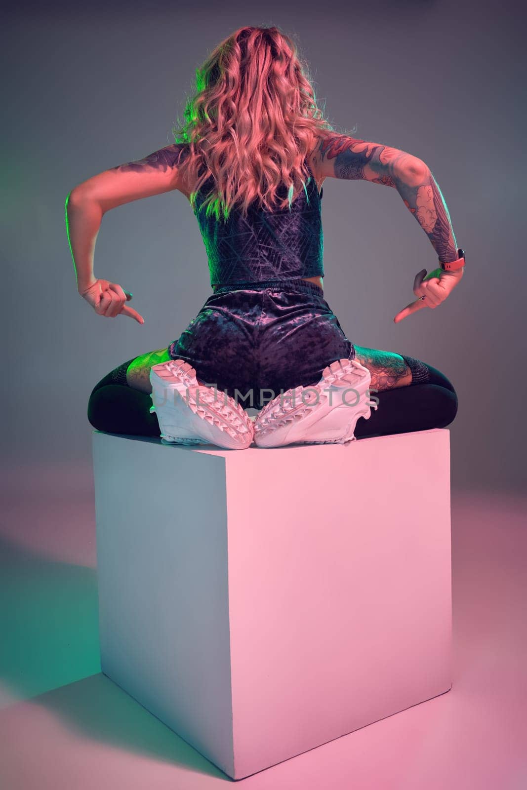 Beautiful young blonde tattoed girl in blue velour booty shorts and t-shirt, she sits on top of big cube, showing off her amazing booty from behind