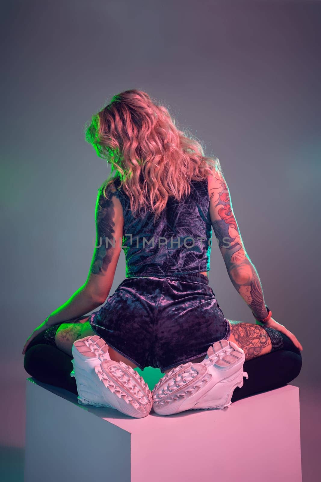 Beautiful young blonde tattoed girl in blue velour booty shorts and t-shirt, she sits on top of big cube, showing off her amazing booty from behind