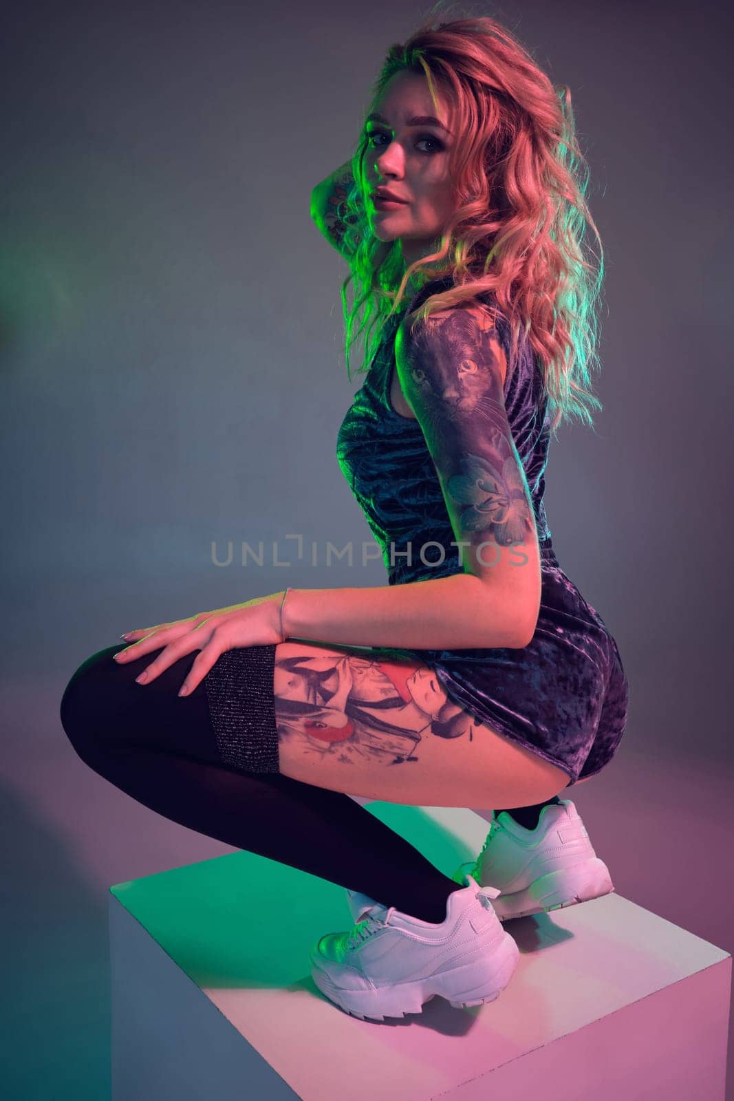 Beautiful young blonde tattoed girl in blue velour booty shorts and t-shirt, making squats on top of big cube, showing off her amazing booty