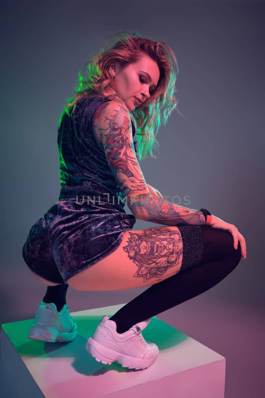 Beautiful young blonde tattoed girl in blue velour booty shorts and t-shirt, making squats on top of big cube, showing off her amazing booty