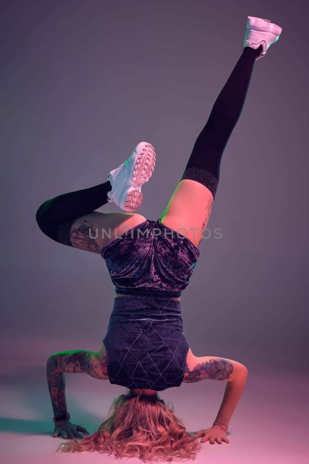 Young beautiful caucasian blonde gymnast woman with tattoo on her body, wearing velour blue booty shorts and black stockings standing upside down, pink background