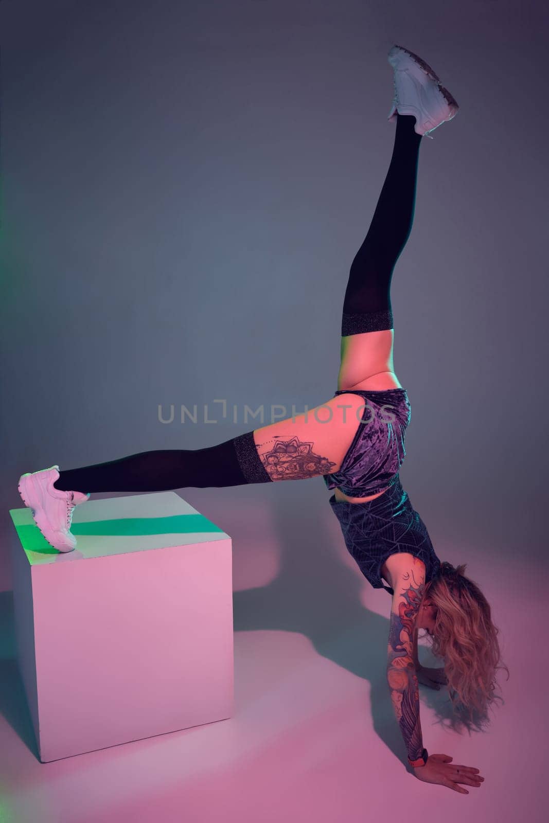 Young beautiful caucasian blonde gymnast woman with tattoo on her body, wearing velour blue booty shorts and black stockings standing upside down using big cube, pink background