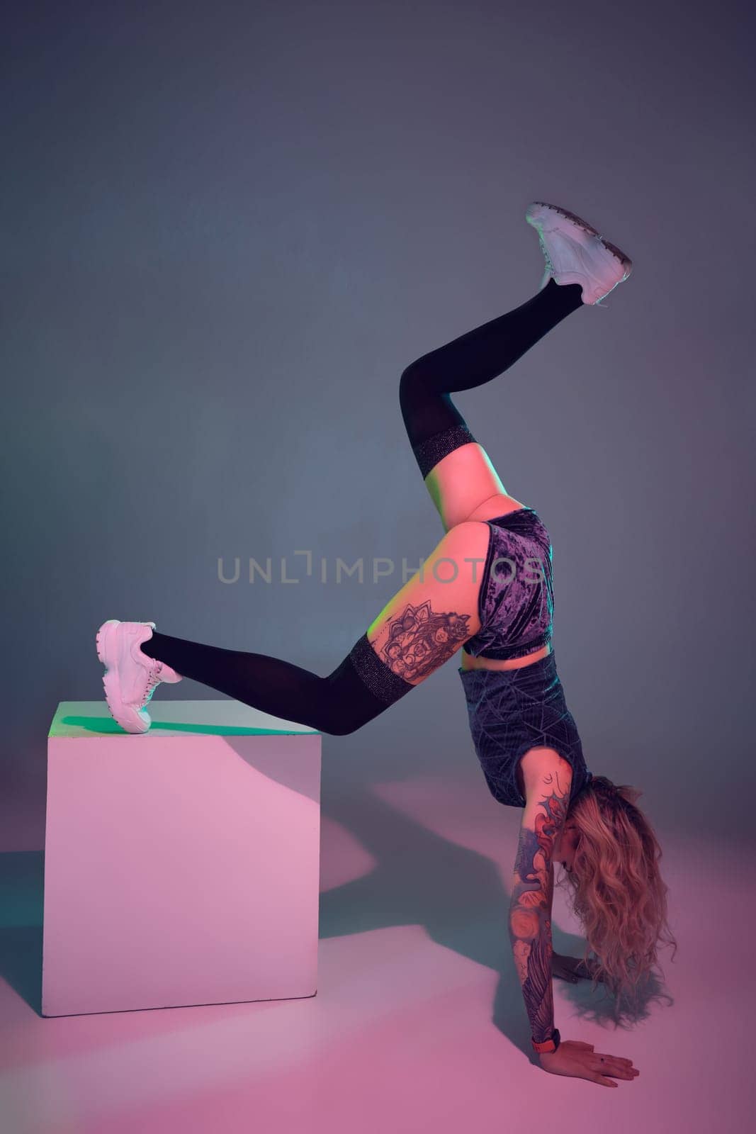 Young beautiful caucasian blonde gymnast woman with tattoo on her body, wearing velour blue booty shorts and black stockings standing upside down using big cube, pink background