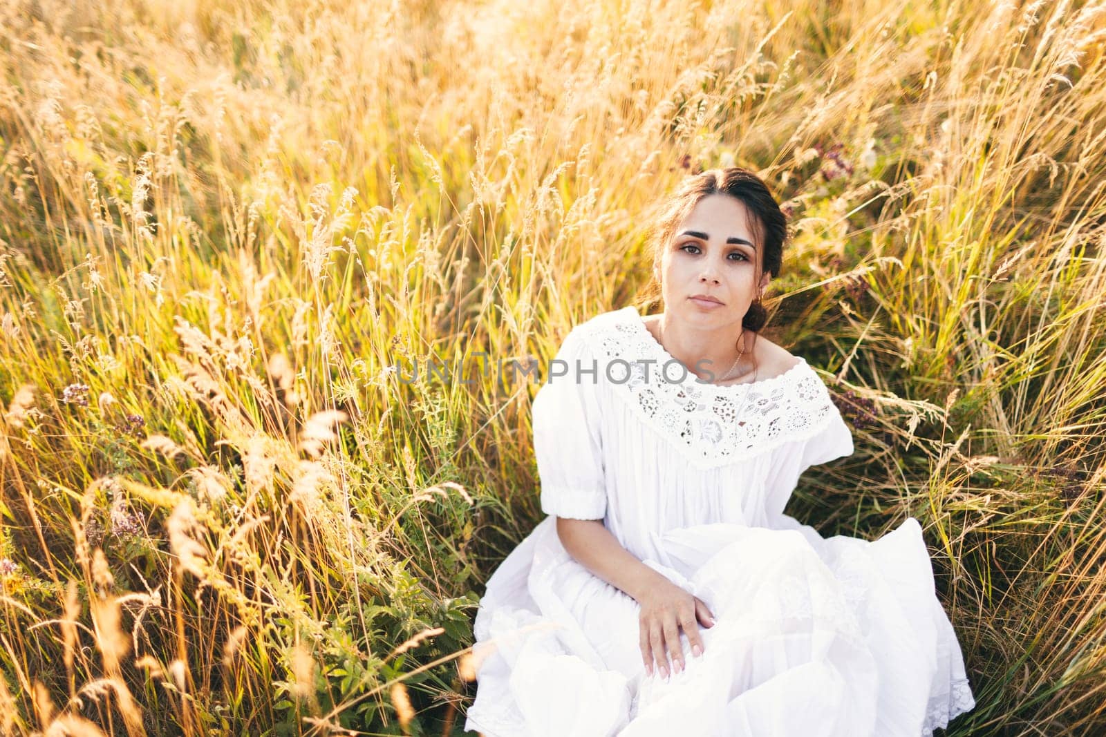Beautiful girl in white dress sitting in wheat field at sunset