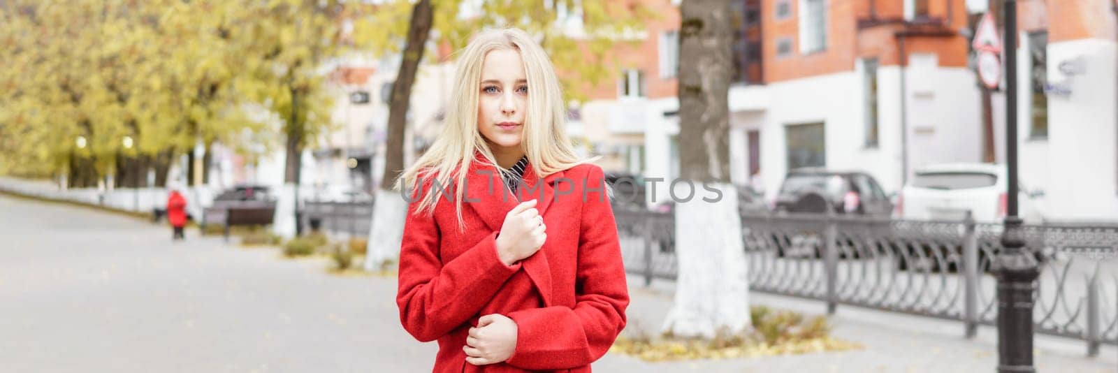 A young blonde woman walks around the autumn city in a red coat. The concept of urban style and lifestyle.