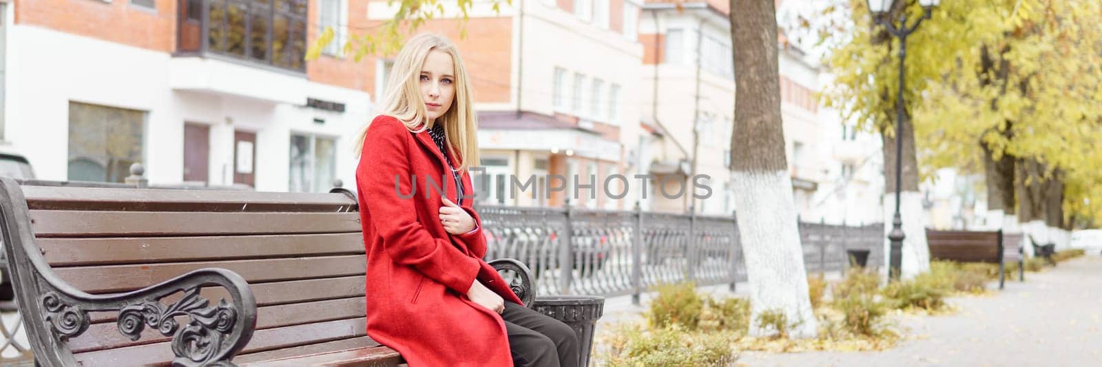 A young blonde woman walks around the autumn city in a red coat. The concept of urban style and lifestyle.