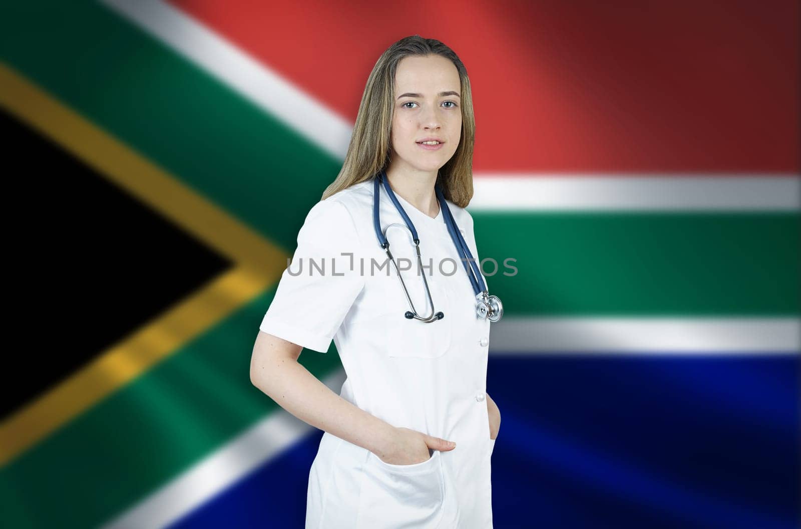 A young female doctor in a white coat and a stethoscope stands on the background of the flag of South Africa.