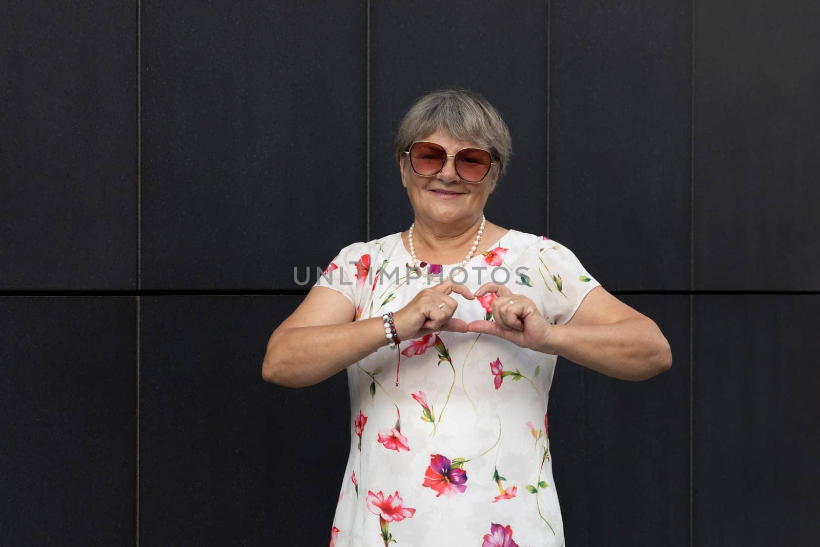 Real Cute Senior Old 70s Woman Makes, Shows Heart Shape With Her Hands and Fingers, Dark Wall on Background. Portrait Of Smiling Kind Elderly Female. Authentic Lifestyle Horizontal High quality photo