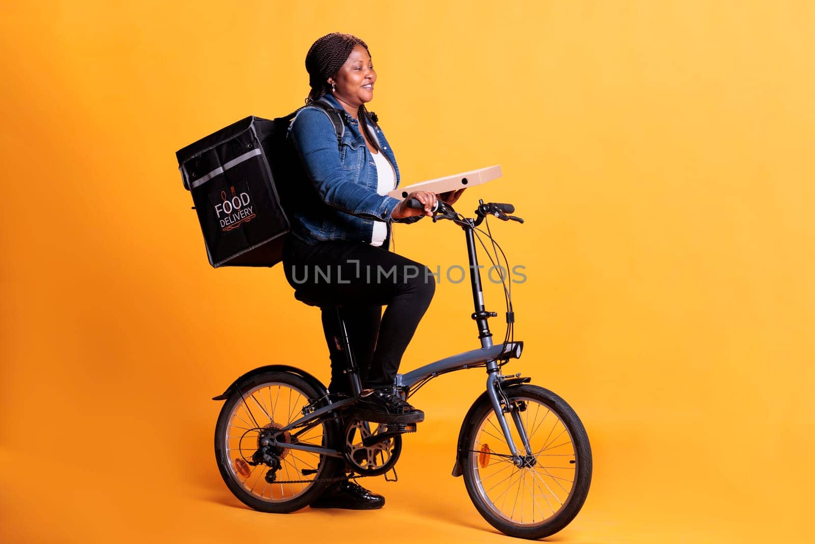 African american pizzeria courier carrying pizza carton box delivering takeaway order to client during lunch time, using bike as transportation in studio. Take out food service and concept