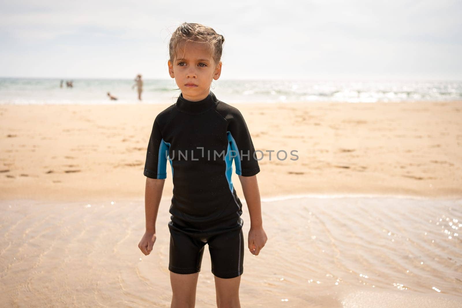 Little girl surfer in wetsuit standing ocean beach. Serious child after surfing on sandy beach