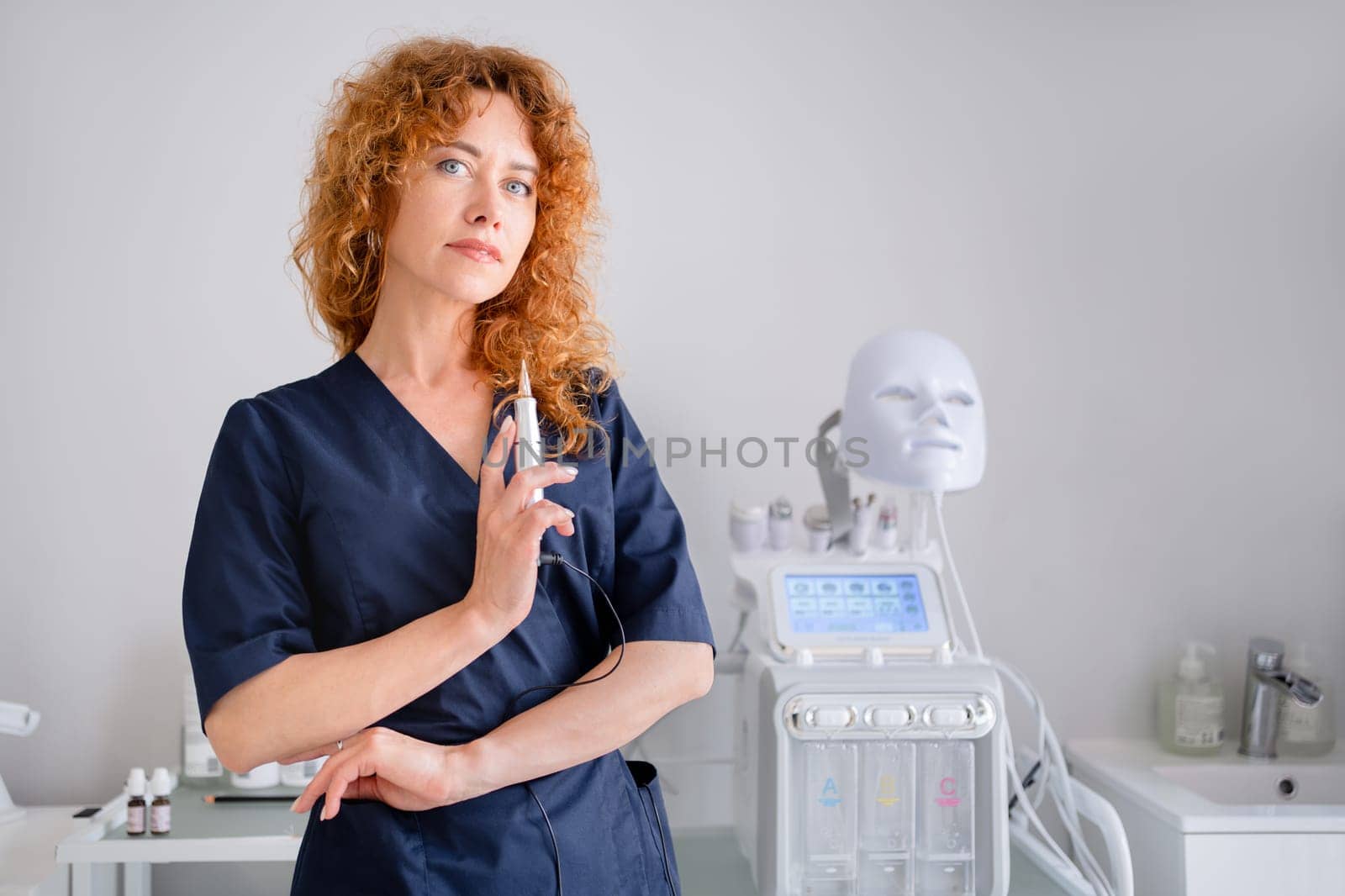Woman cosmetologist standing hold device and looking at camera in beauty spa salon. Female dermatologist, skin therapist, beautician skincare professional in clean white coat uniform