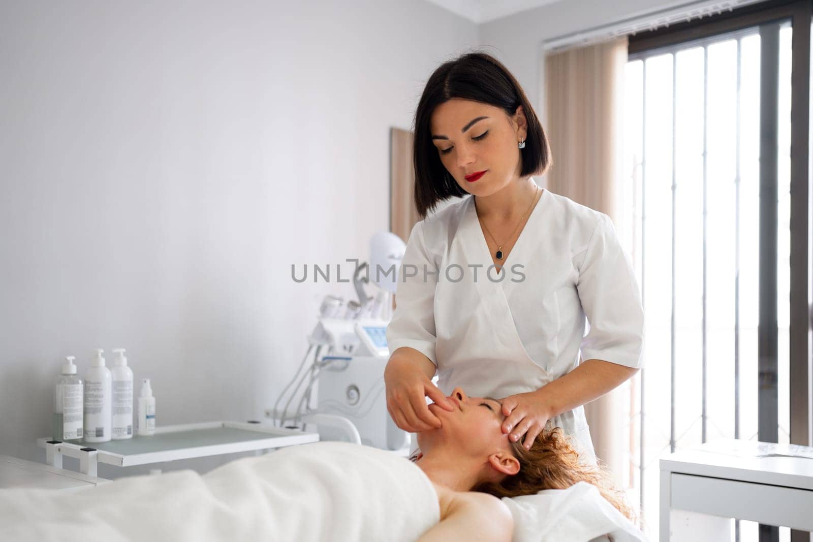 Massage therapist providing facial massage to woman at cosmetology clinic. Beautician administering skincare and stimulation procedures. Beauty salon offering spa treatments and massages.