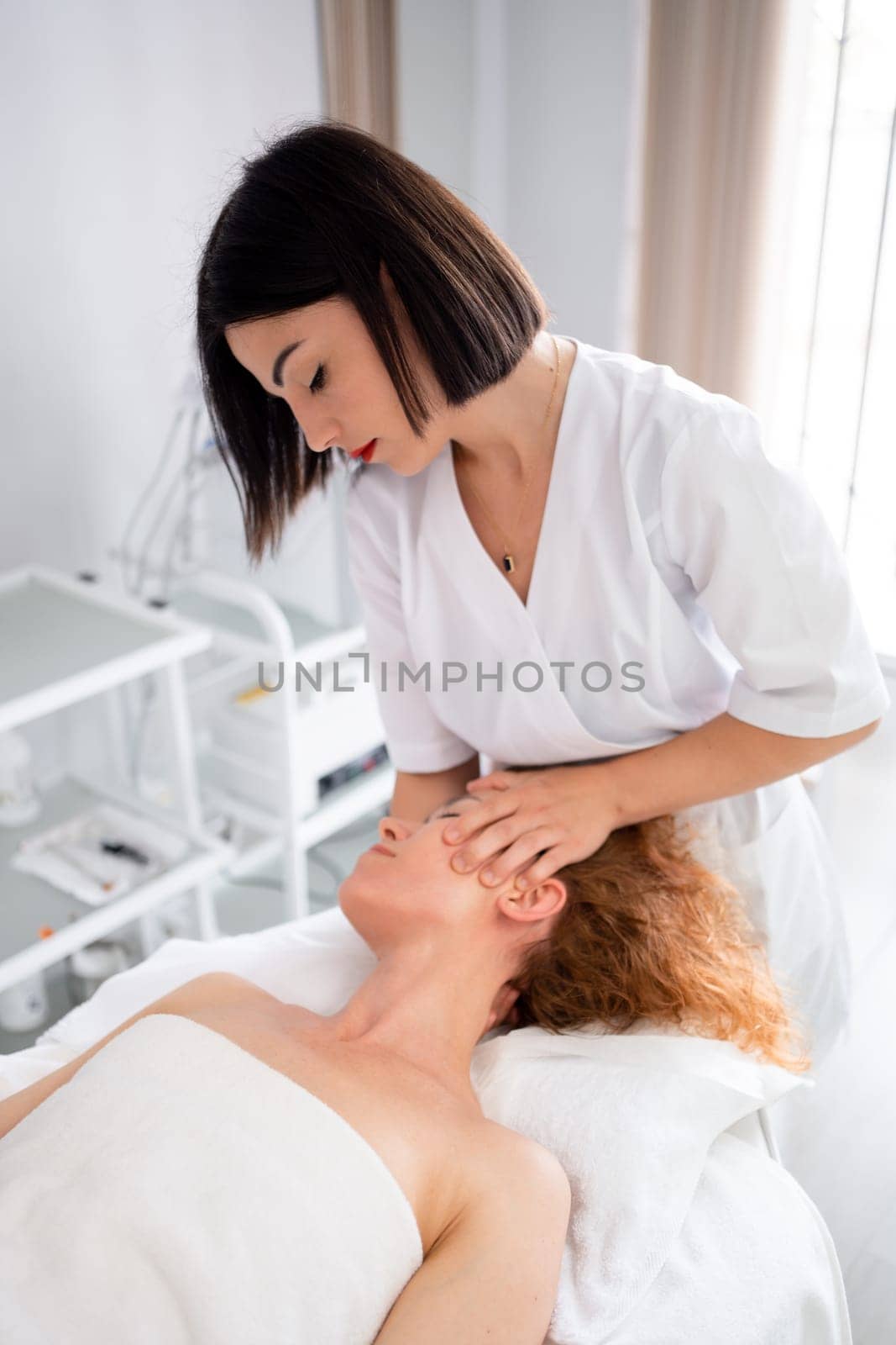 Massage therapist massaging head, shoulders and neck to woman in cosmetology clinic. Beautician making skincare and stimulation procedure. Spa treatment and massage in beauty salon. Vertical photo