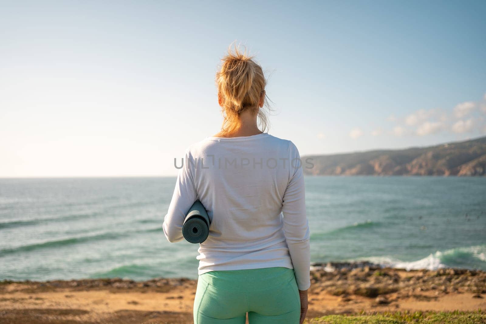 Back view senior woman holding yoga mat on ocean shore. Retired woman enjoying ocean views before practicing yoga. Feeling harmony. Healthy healthy senior lifestyle concept