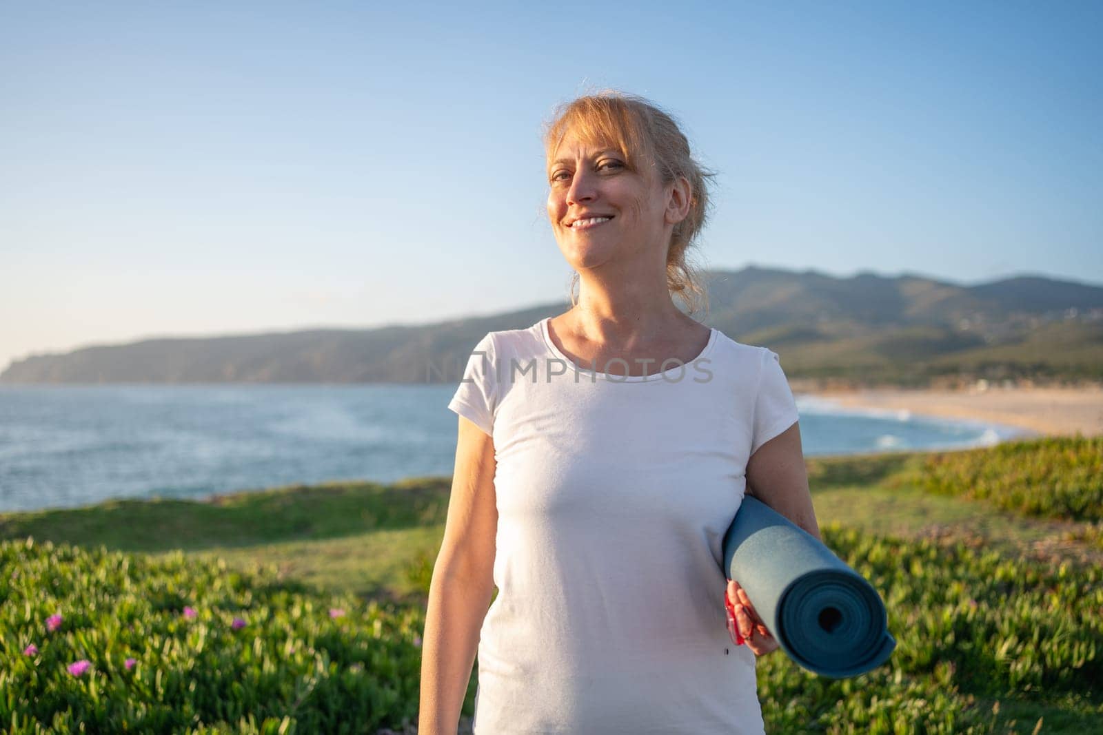 Elderly Caucasian lady holding yoga mat smiling on beach. Getting ready for morning yoga session on ocean shore, promoting healthy lifestyle and idea of enjoying retirement on vacation