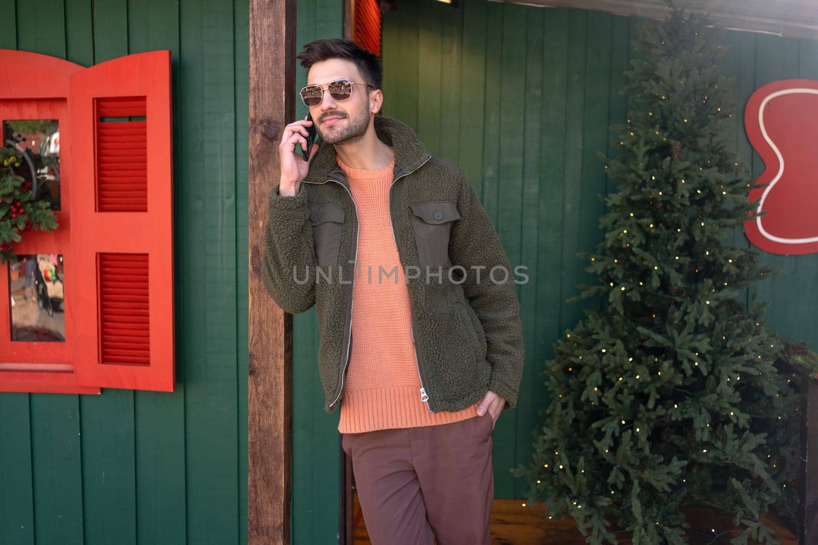 Handsome Caucasian man wearing sunglasses standing decorated town near Christmas tree while talking on his smartphone. He appears stylish as he extends holiday greetings using his mobile phone