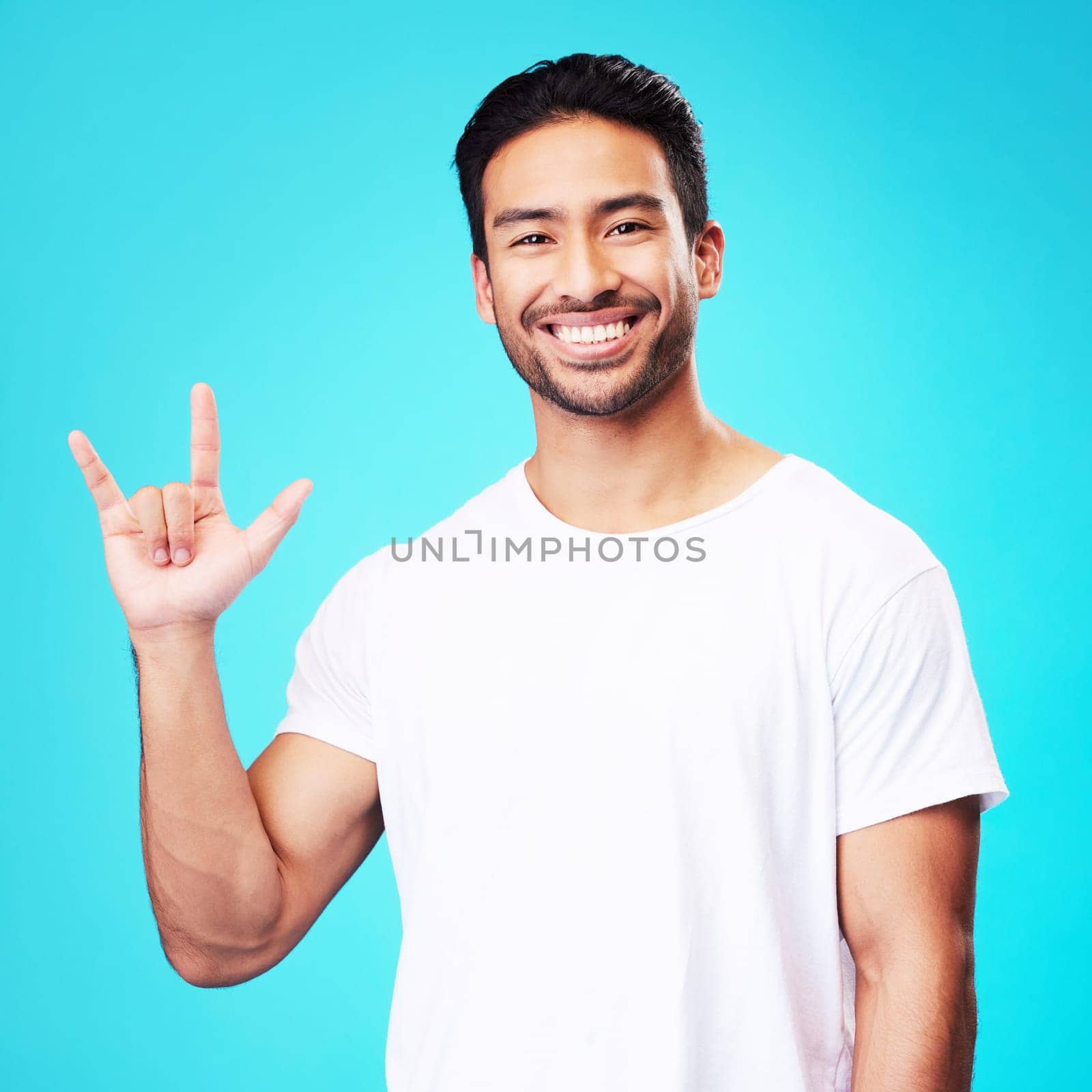 Man, horns and love in studio portrait with smile, kindness and sign language by blue background. Young guy, student and happy with icon, emoji and hand with care, romance or vote with white t-shirt.