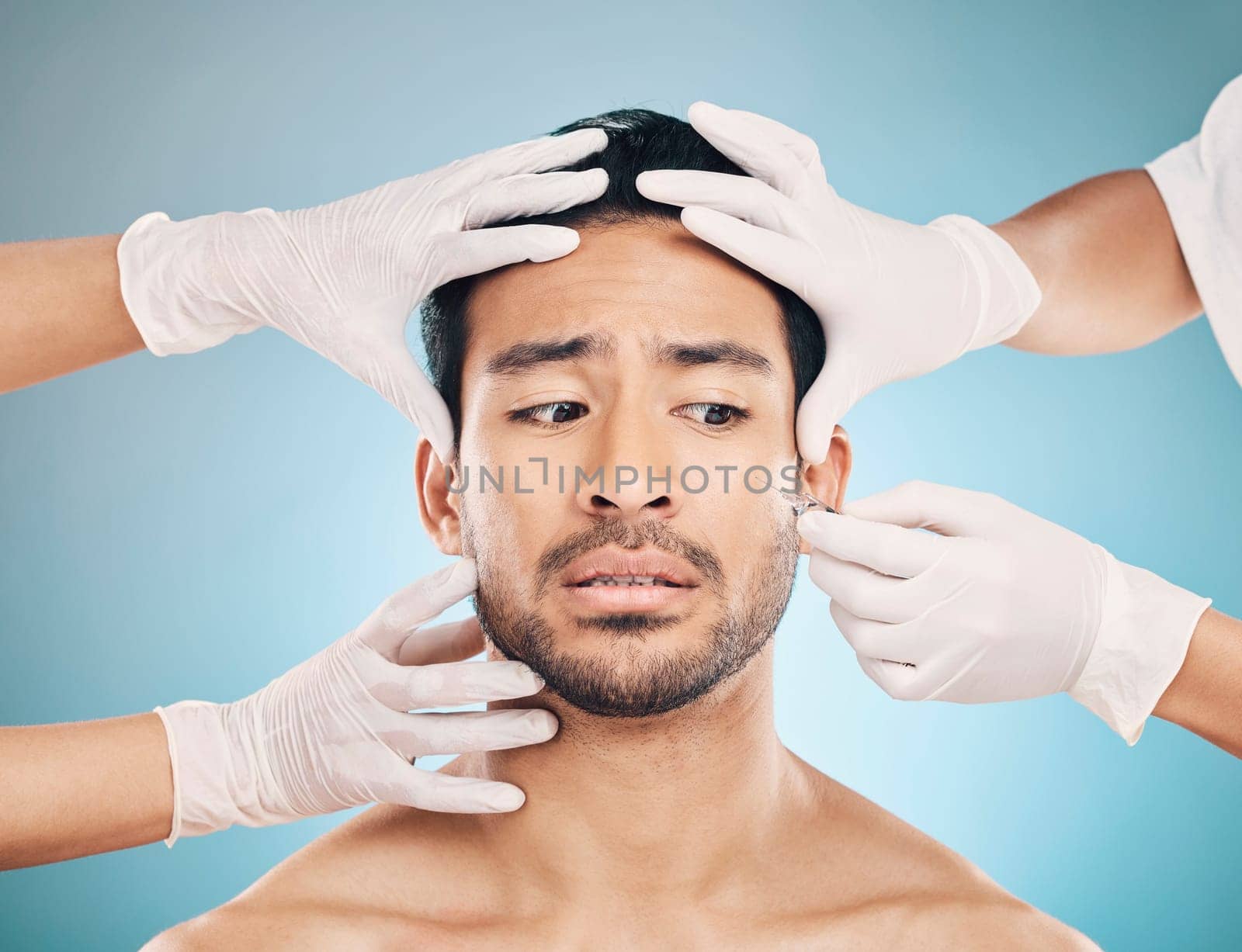 Face, hands and plastic surgery with a worried man in studio on a blue background for beauty enhancement. Aesthetic, botox or change with a young male patient looking nervous in a clinic for skincare by YuriArcurs