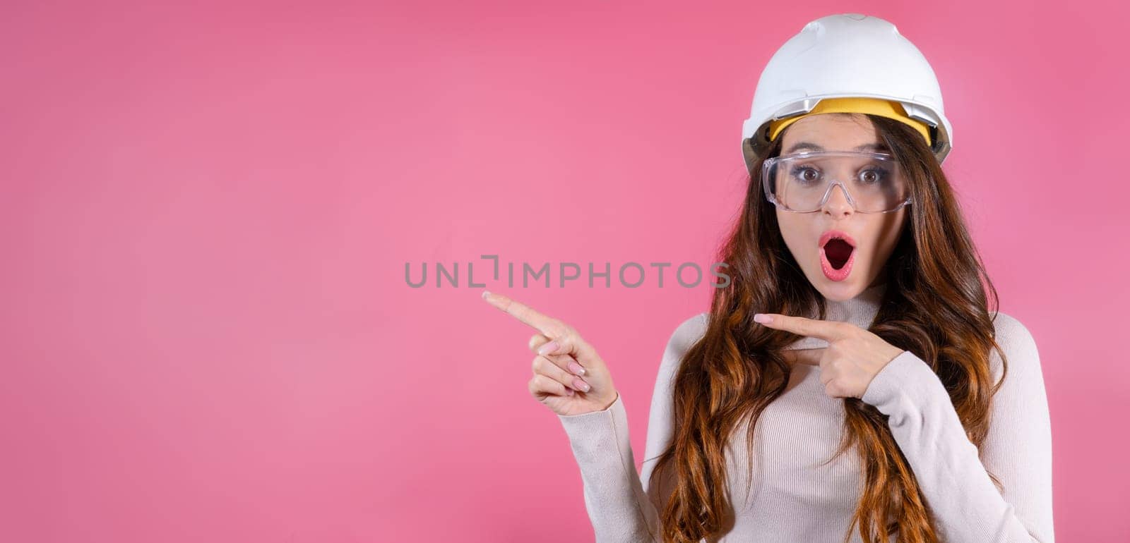 A joyful woman architect in helmet celebrating the successful completion of her project and shows on the copy space on the pink background