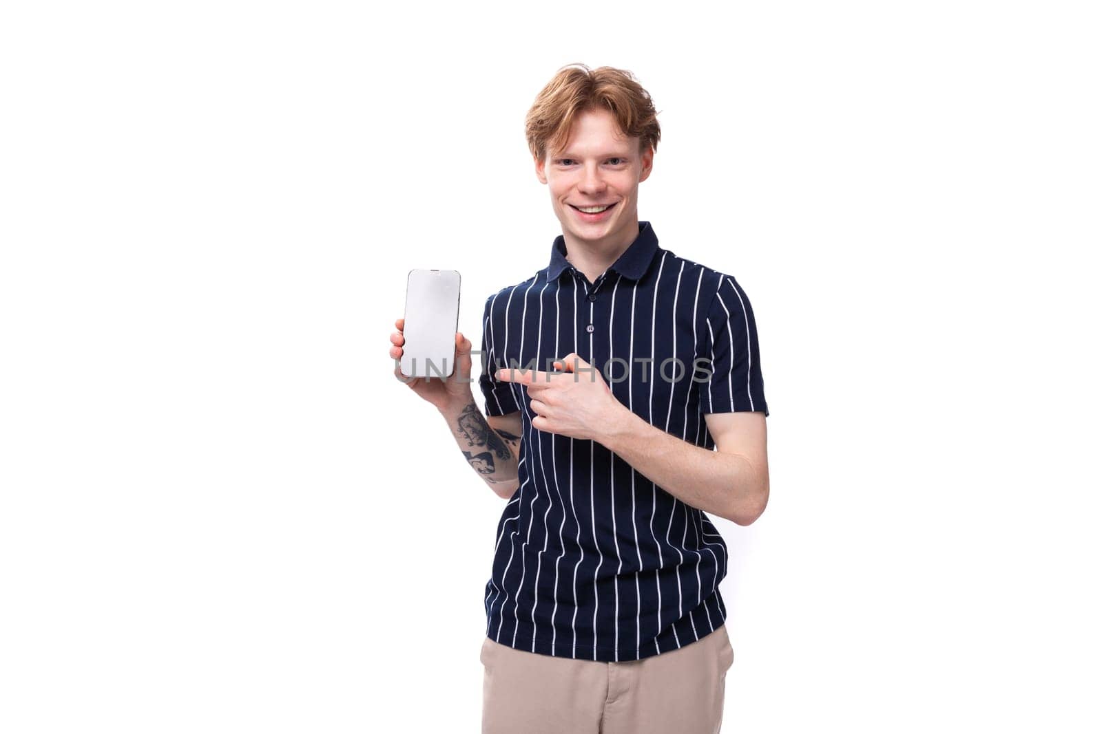 joyful stylish young blond guy in a striped polo shirt with a tattoo shows a phone mockup on a white background.