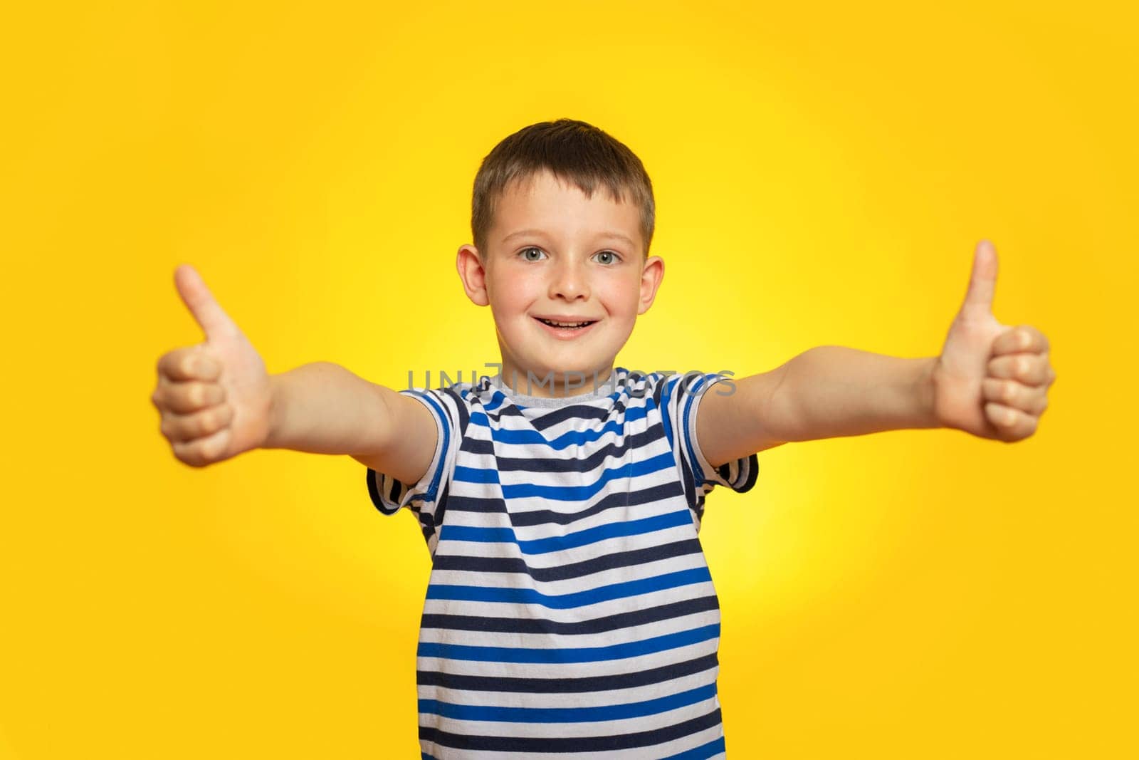 Portrait of happy boy in striped t-shirt showing thumbs up gesture on yellow background.