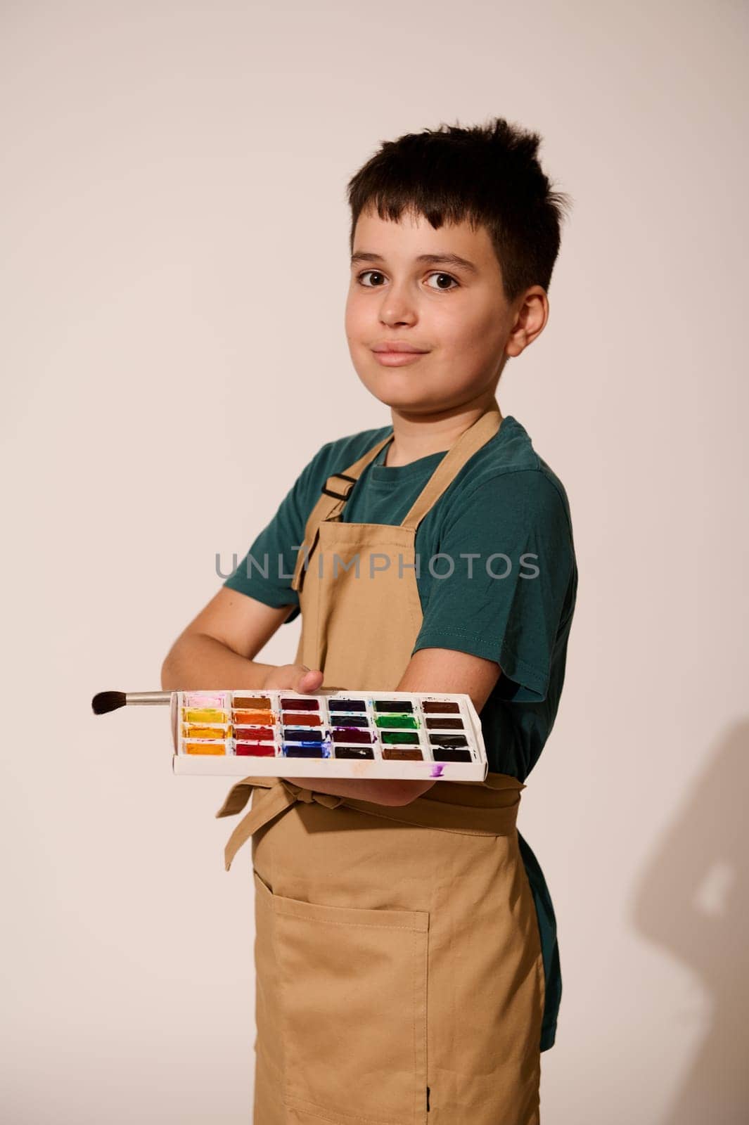 Vertical portrait of adorable teenage boy wearing beige apron, holding paintbrush and palette with watercolors, smiling, looking at camera, standing with folded hands over isolated studio background