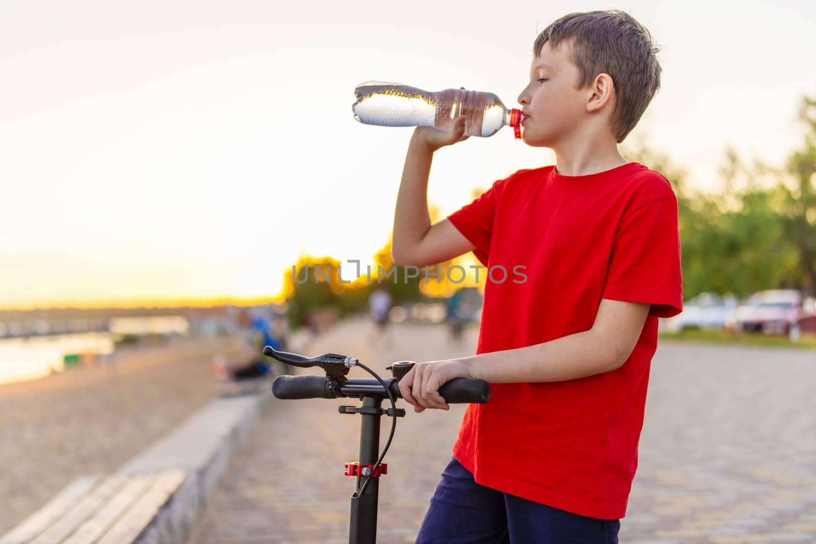 A boy drinks water from plastic bottle, standing with scooter.