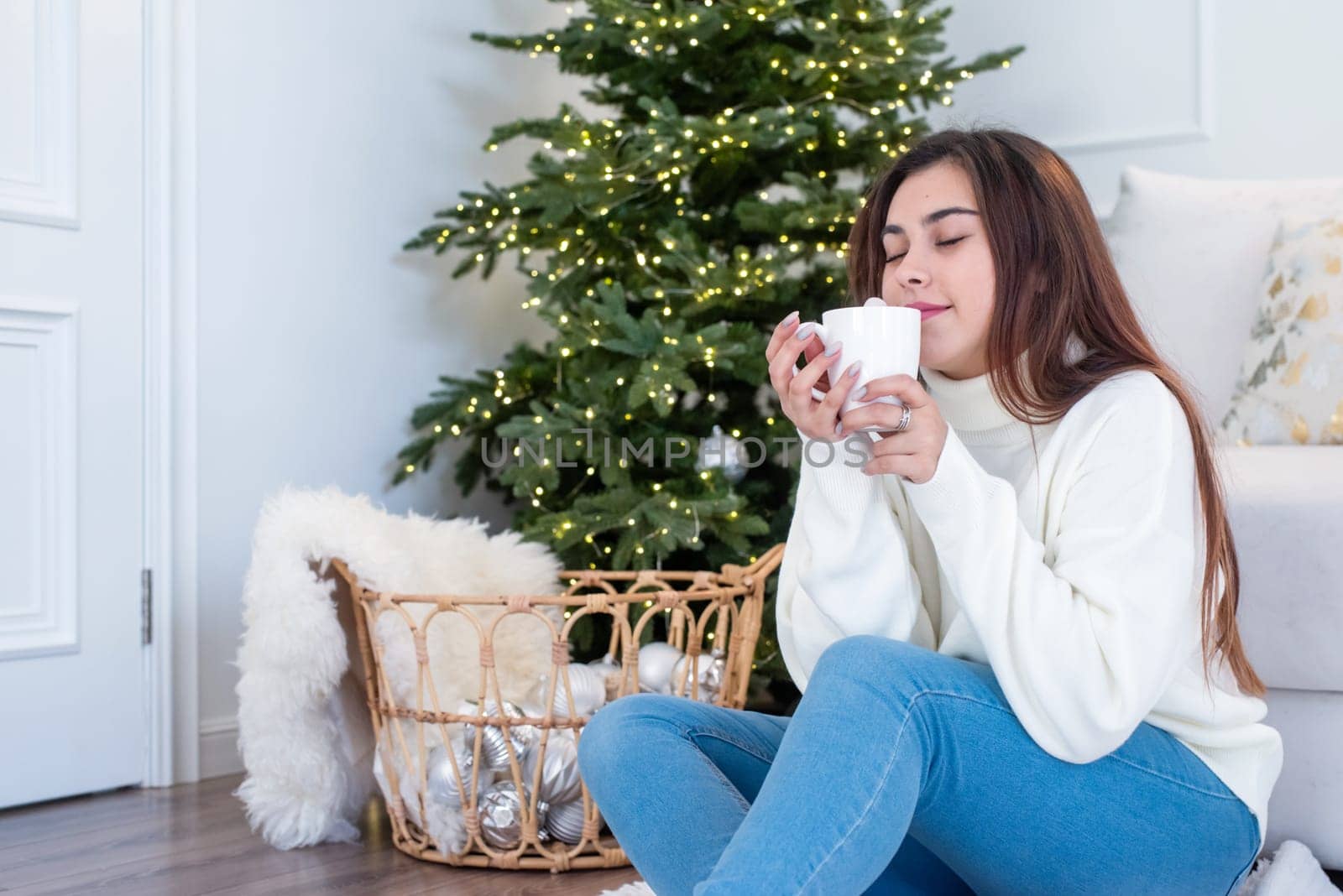 Woman in warm white winter sweater sitting at windowsill at home at christmas eve holding cup with marshmallows by Desperada