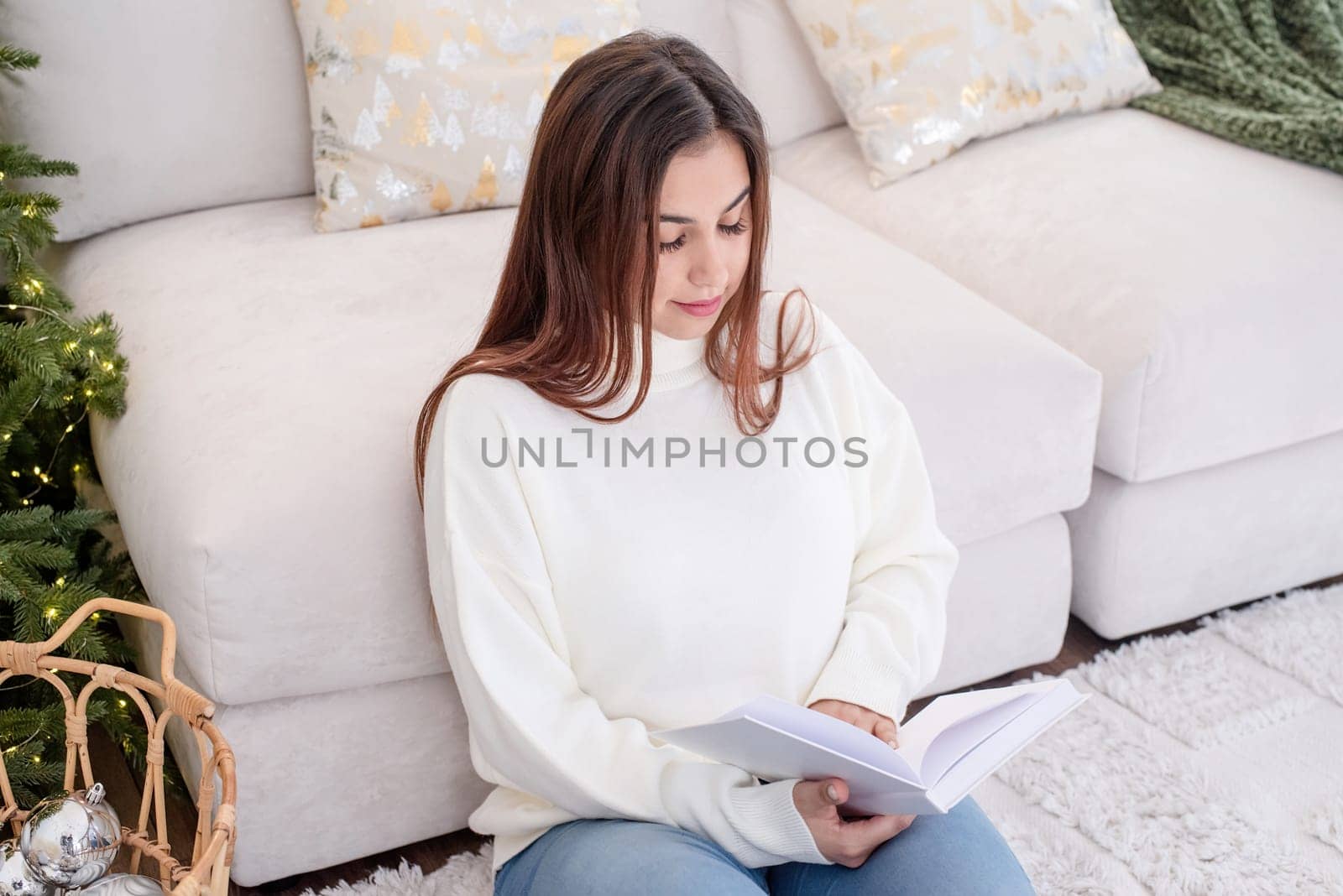 Merry Christmas and Happy New Year. Woman in warm white winter sweater sitting on couch reading a book