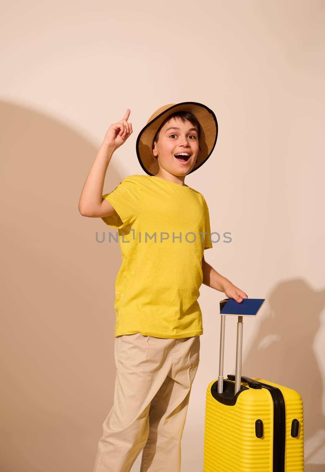 Traveler teen boy in straw hat, with suitcase and passport, points finger aside a copy ad space, expressing idea and sharing happy positive emotions, looking at camera, isolated over studio background