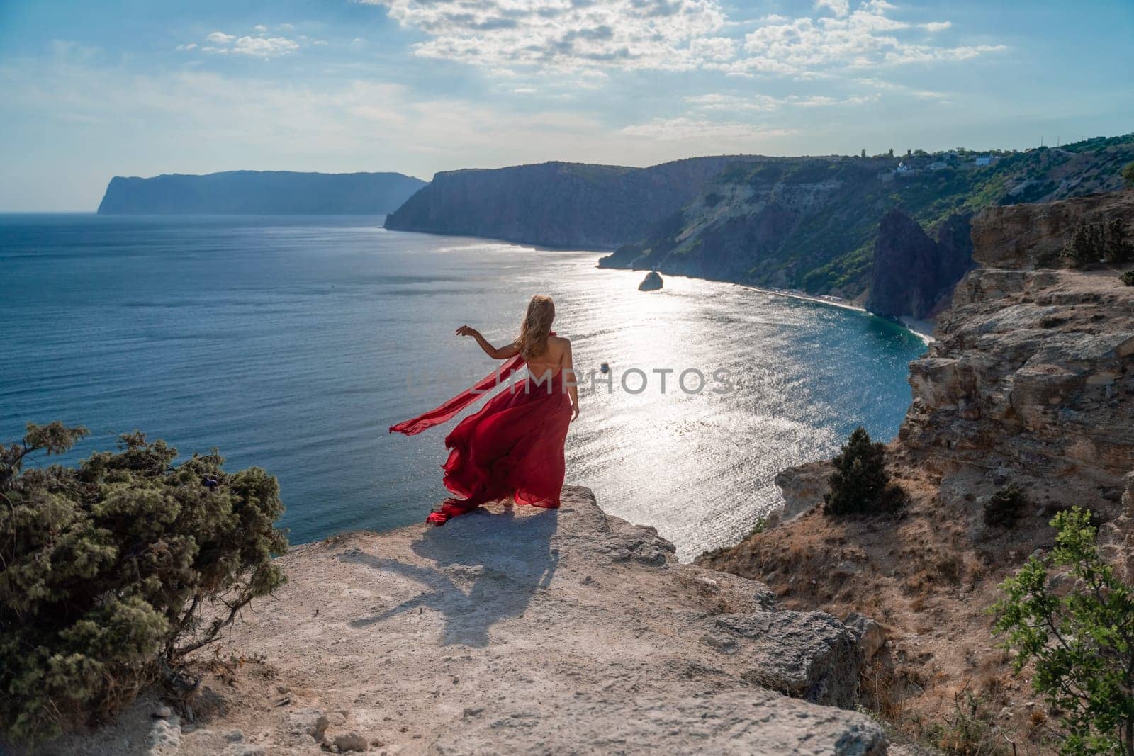 A girl with flowing hair in a long red dress stands on a rock above the sea. The stone can be seen in the sea. Sunny path to the sea from the sun