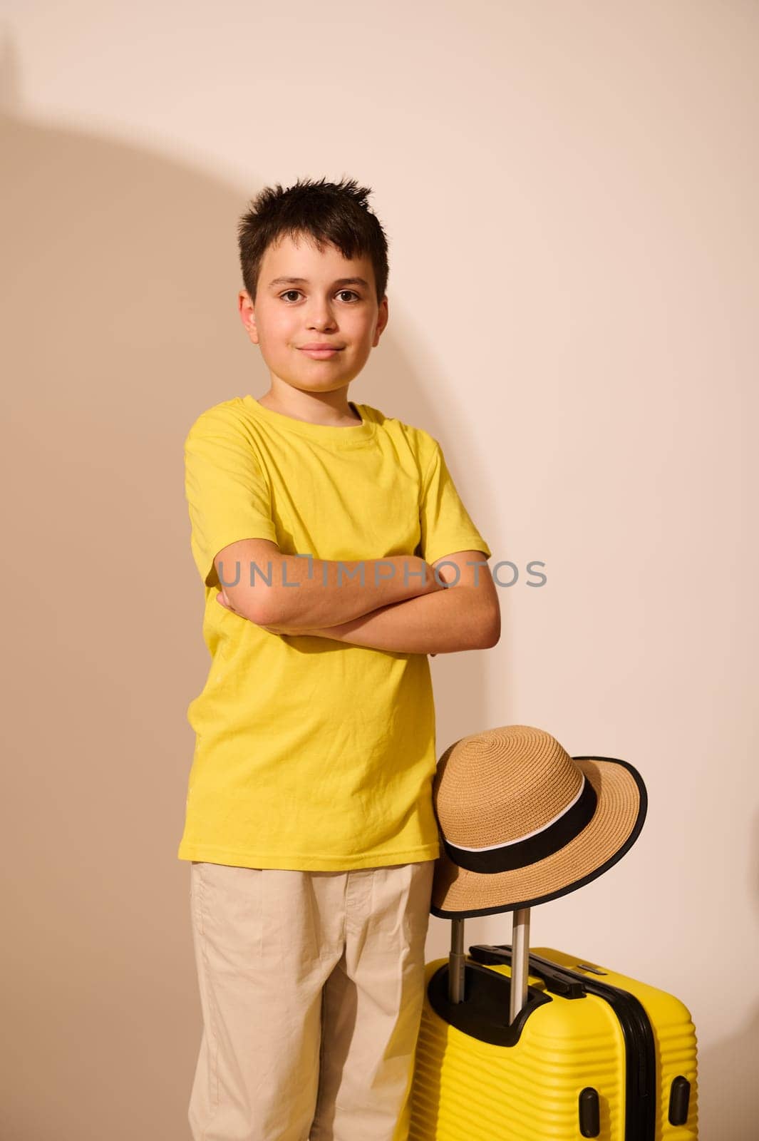 Confident portrait of a handsome multiethnic teenager in yellow t-shirt and gray trousers, crossing his arms, traveling abroad, standing with his yellow suitcase, isolated on white studio background