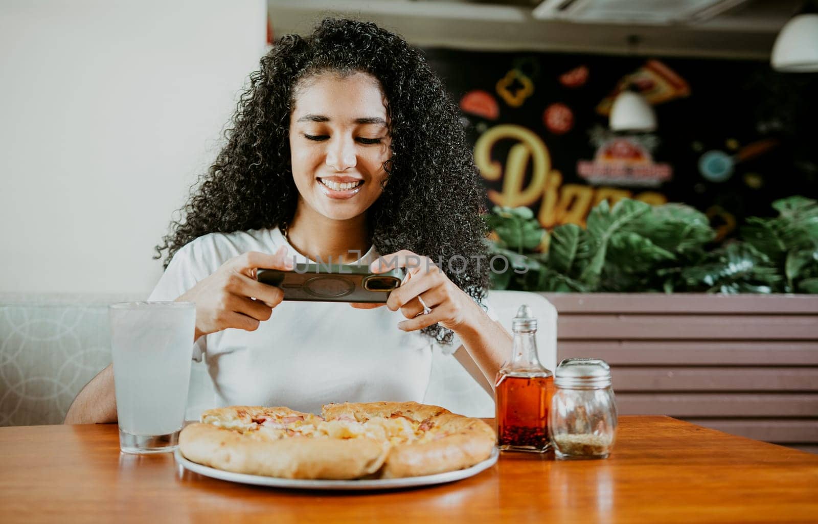 Smiling young woman using phone photographing a pizza in a restaurant. Happy girl photographing a pizza with the cell phone by isaiphoto