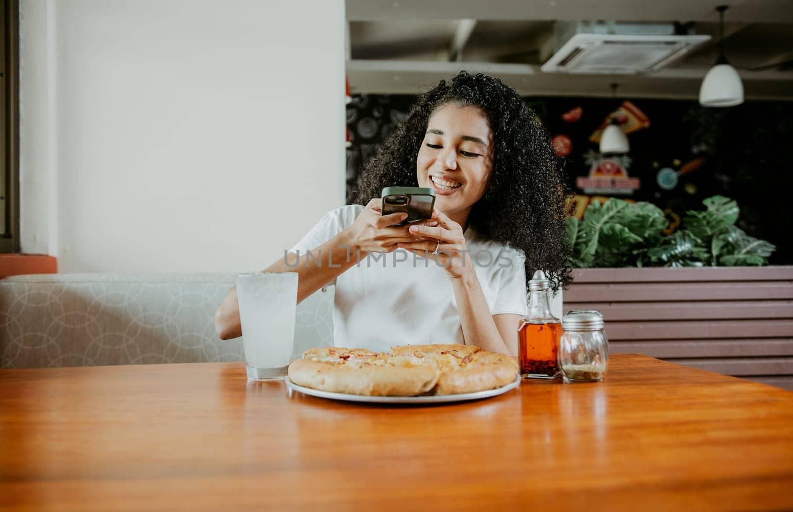 Afro girl taking a picture of a pizza with the phone in a pizzeria. Smiling young woman using phone photographing a pizza in a restaurant by isaiphoto