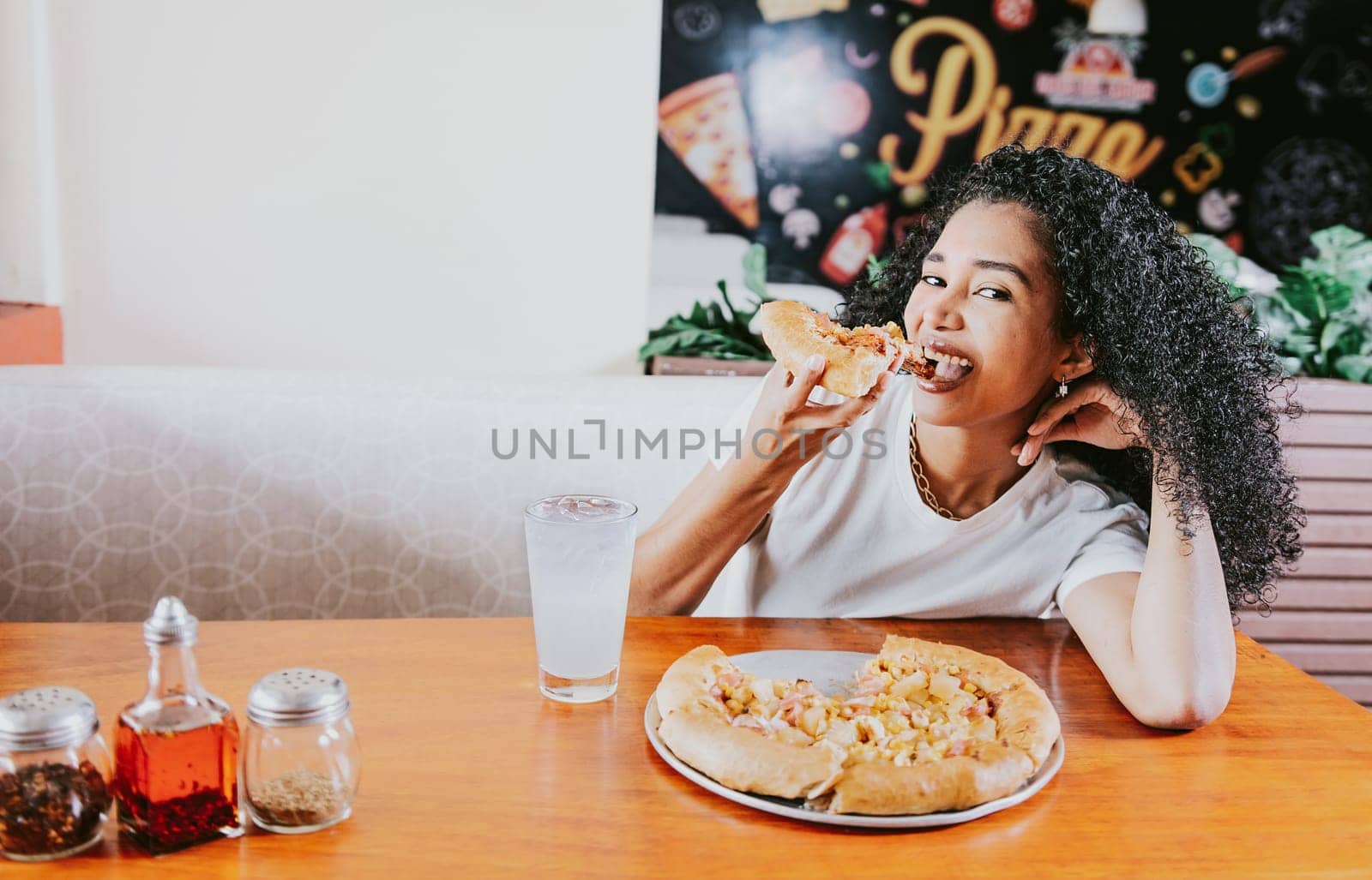 Smiling young woman enjoying a pizza in a restaurant. Happy afro hair girl eating pizza in a restaurant, eating and looking at camera