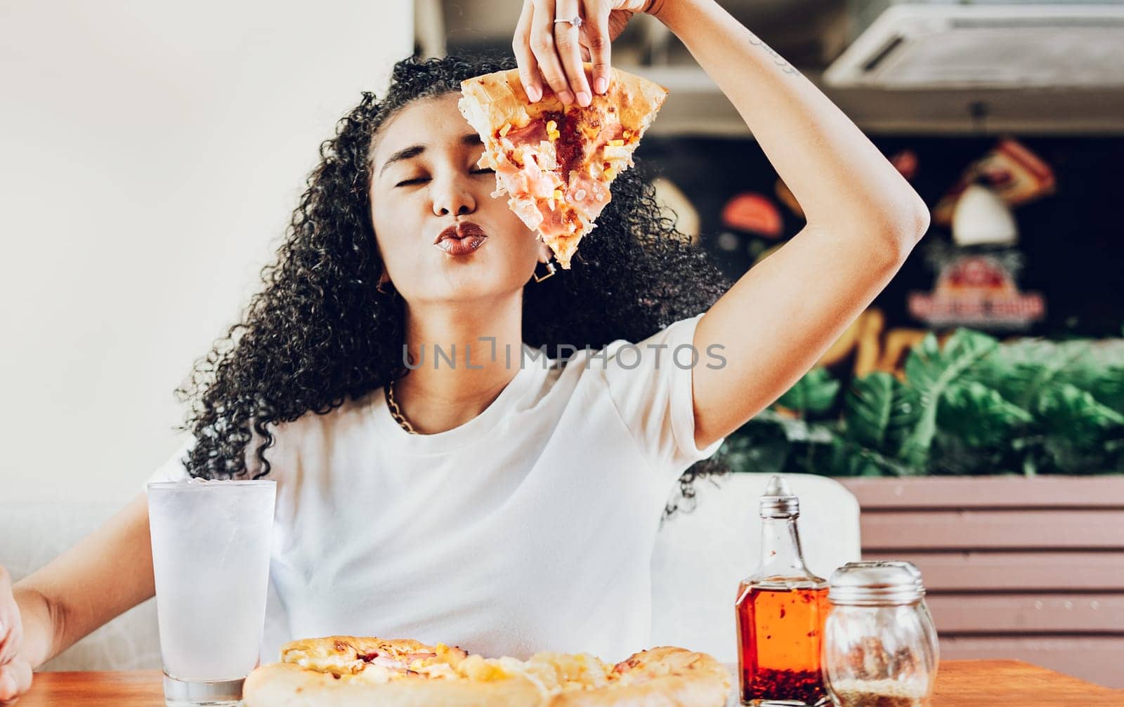 Girl in a restaurant showing slice of pizza giving a kiss to the camera. Beautiful curly haired woman showing slice of pizza in a restaurant with copy space