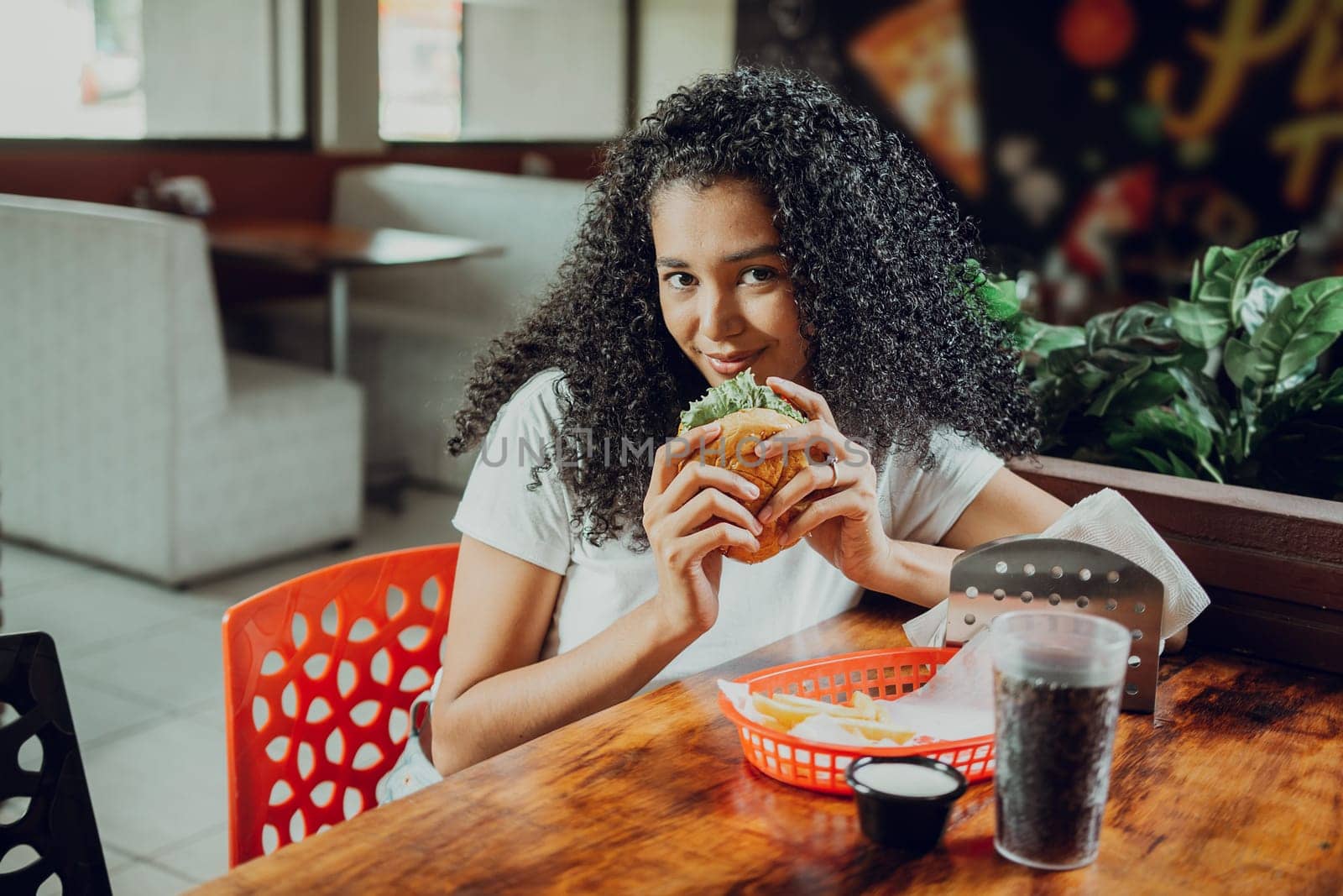 Portrait of afro girl holding a hamburger in a restaurant. Beautiful latin girl enjoying a delicious hamburger in a restaurant
