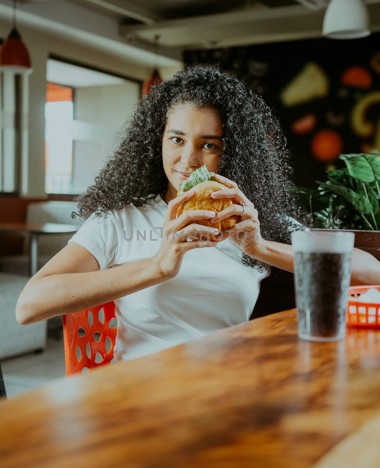 Beautiful latin girl enjoying a delicious hamburger in a restaurant. Portrait of afro girl holding a hamburger in a restaurant.