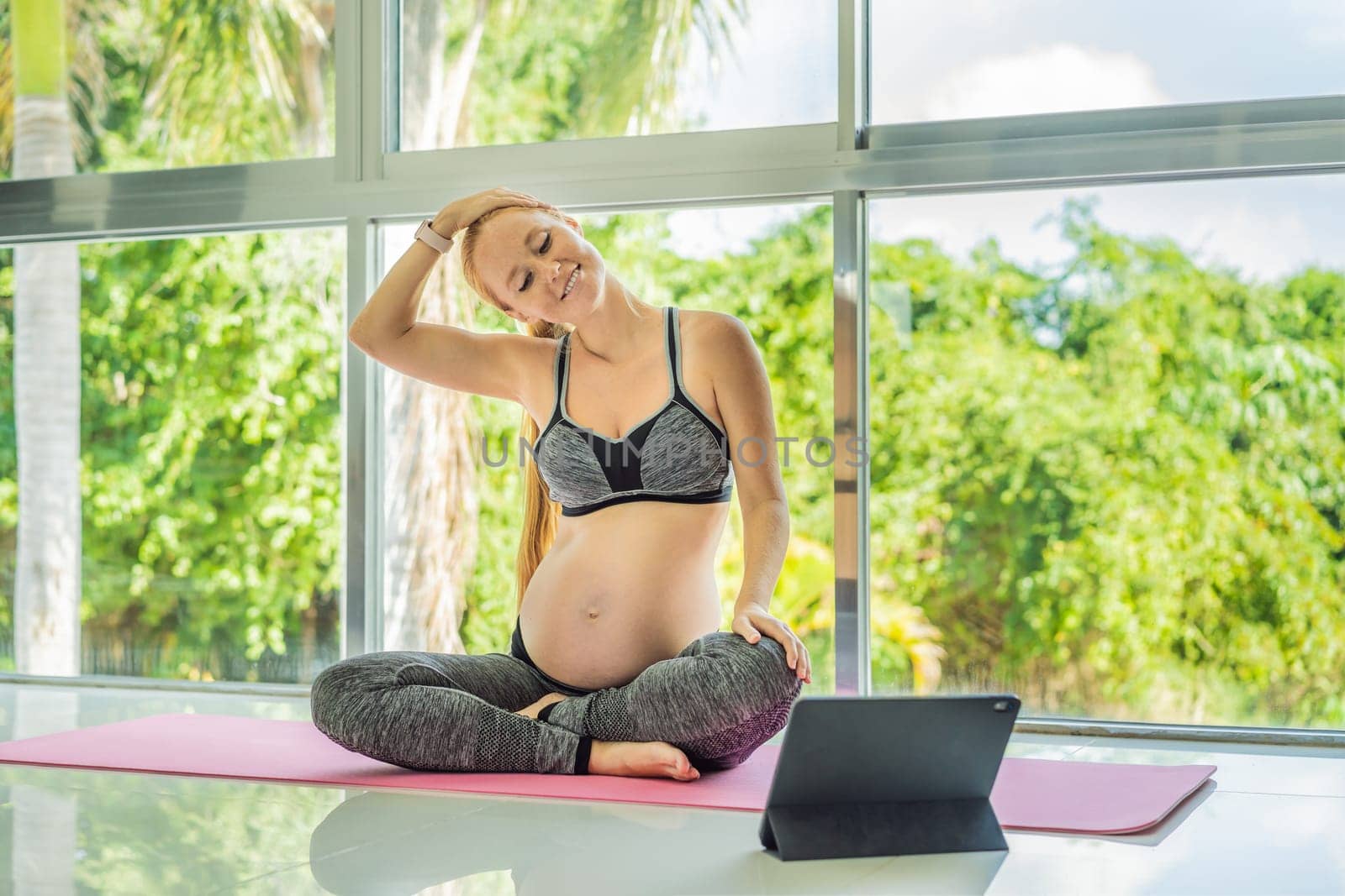 Portrait of young pregnant woman doing yoga on mat with online video lessons indoors on a daytime. Balance exercises. Motherhood, health care, sportive lifestyle, fitness, wellness, pregnancy concept.