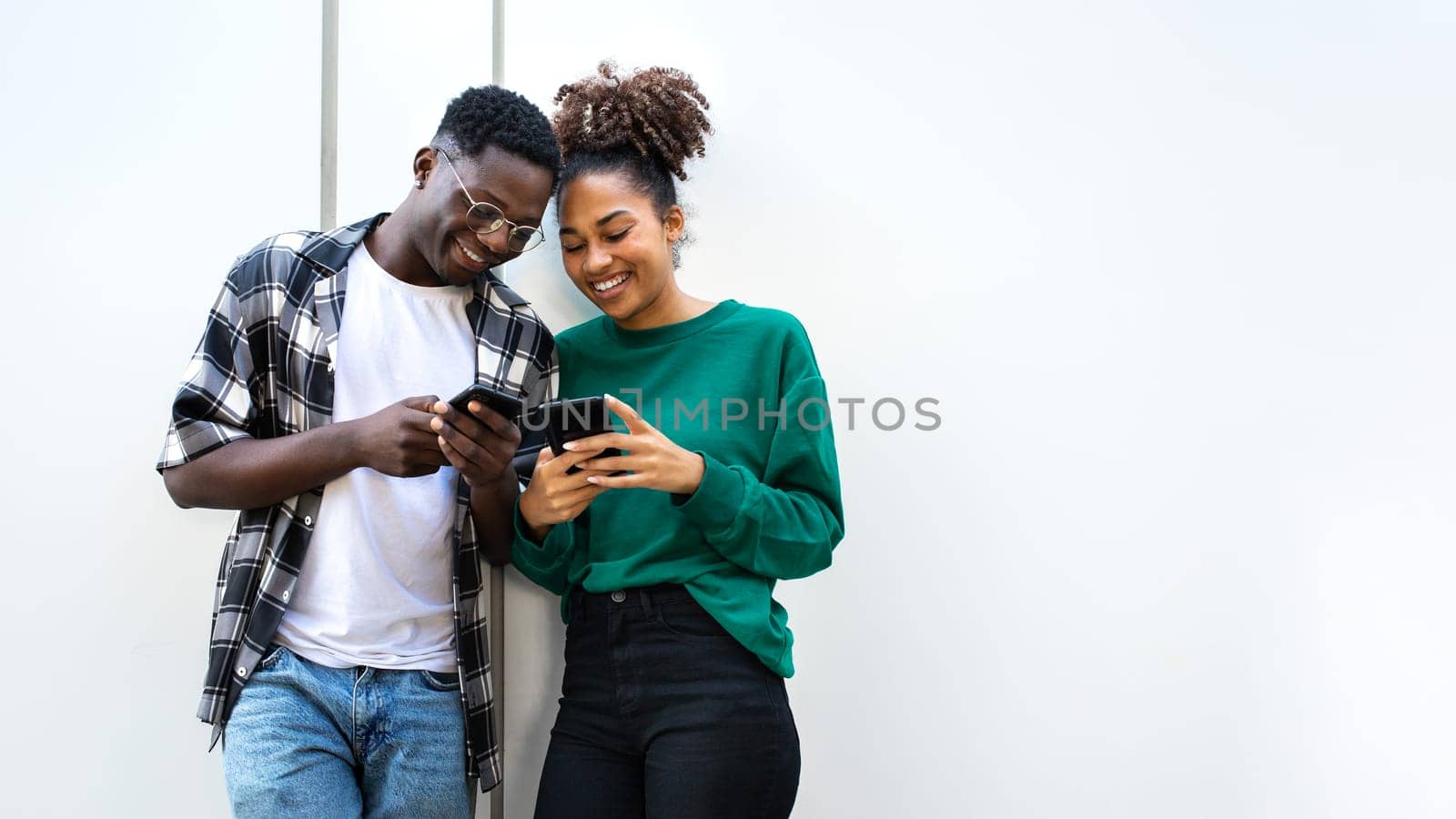 African American young couple looking smart phones together standing against white wall. Copy space. Technology.