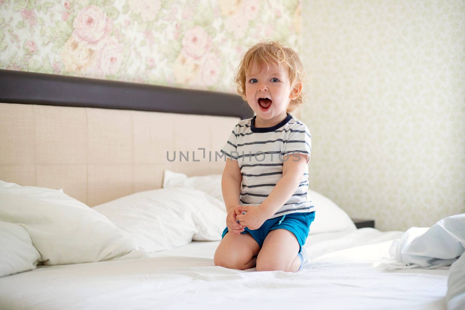 Cute laughing child boy sits in bed. Boy plays at home. Child in a sunny bedroom.