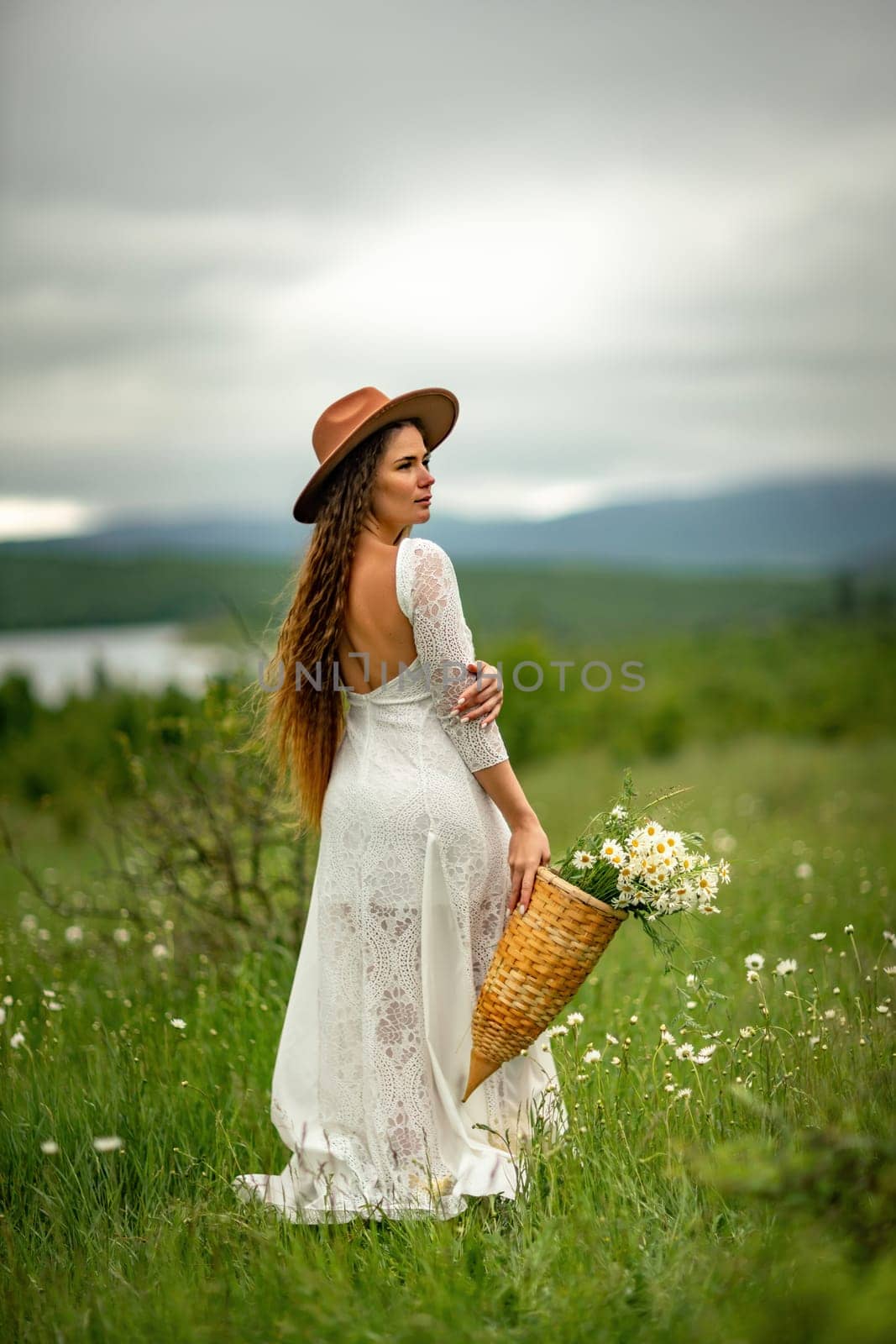 A middle-aged woman in a white dress and brown hat stands on a green field and holds a basket in her hands with a large bouquet of daisies. In the background there are mountains and a lake. by Matiunina