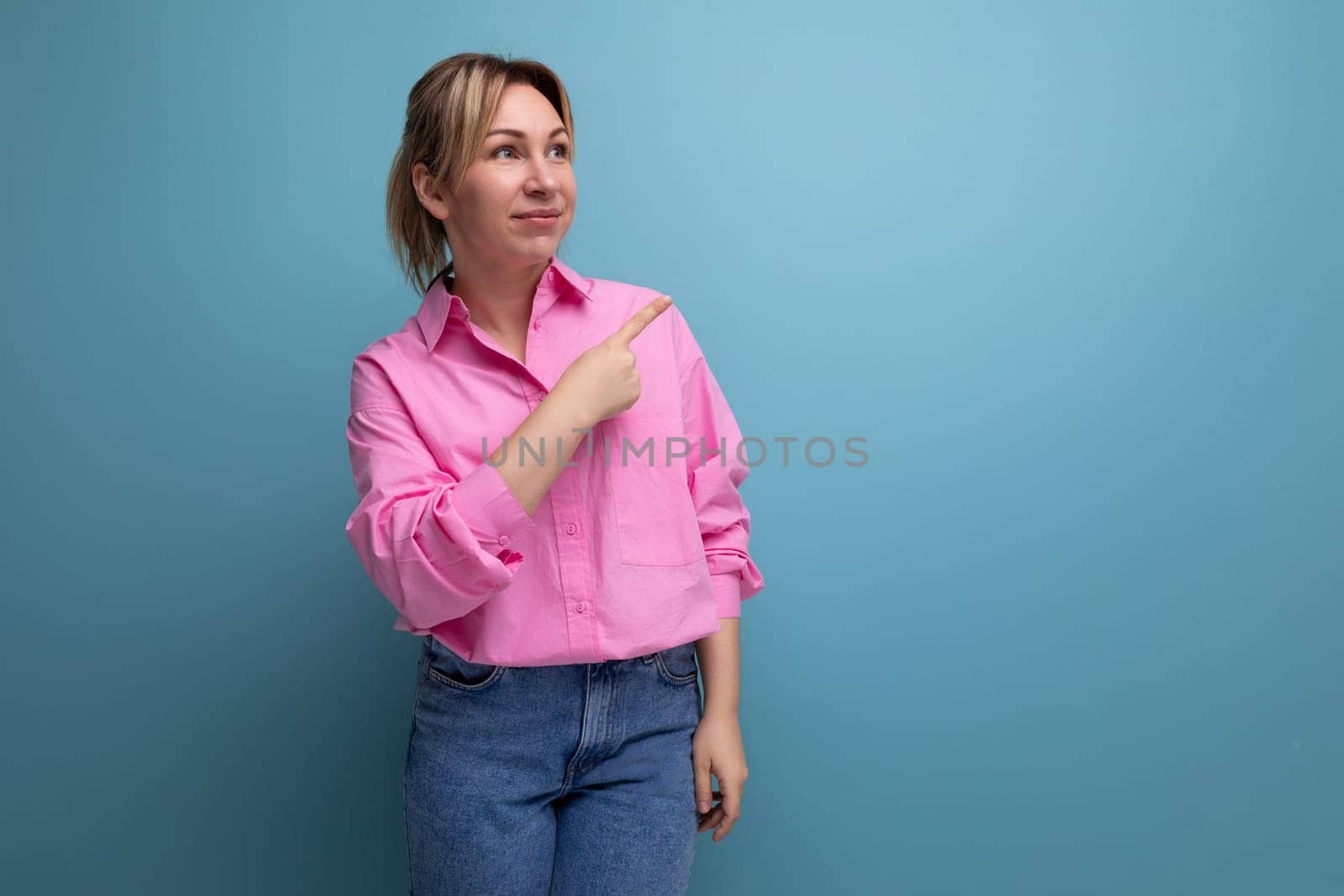 young well-groomed pretty european blonde secretary woman dressed in a pink shirt shows her hand to the side of an advertisement offer on a studio background with copy space by TRMK
