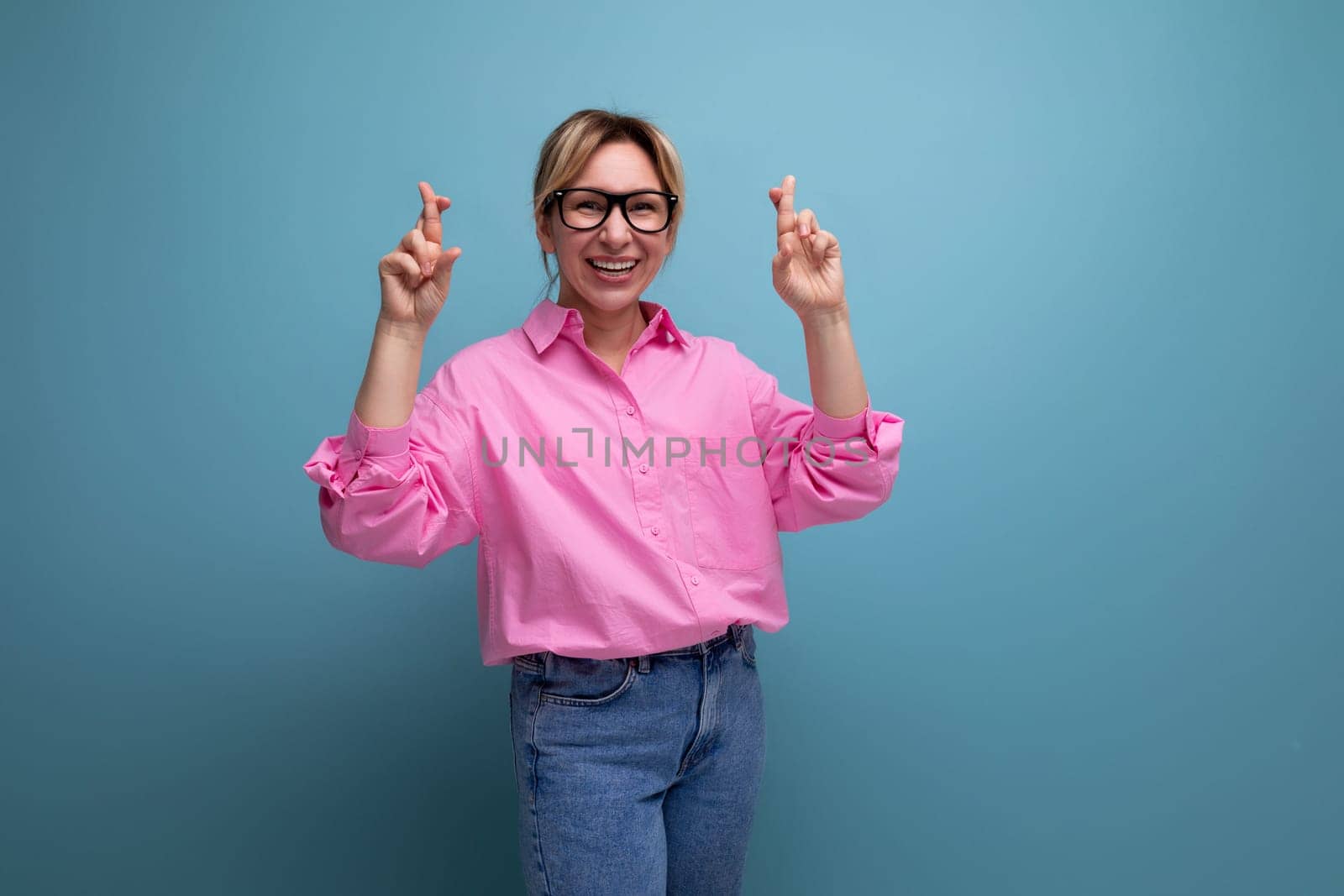 young caucasian blonde secretary woman with ponytail hairstyle dressed in pink shirt crossed her fingers on studio background with copy space.