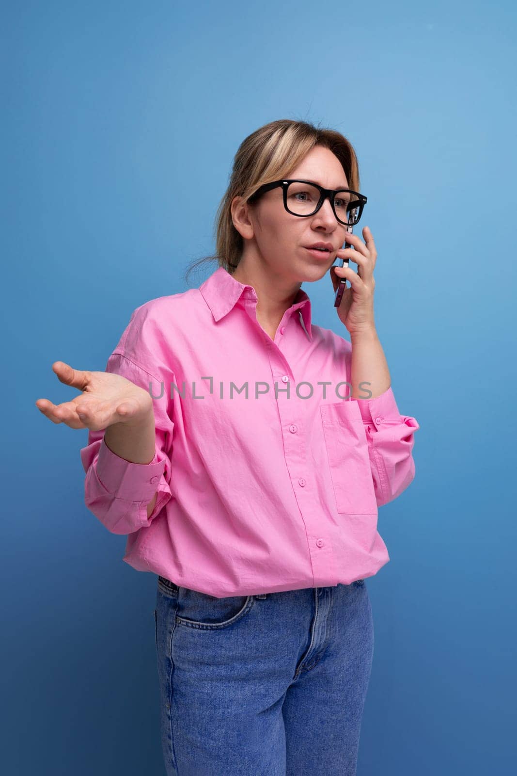 young confident blond woman with ponytail and glasses dressed in a fashionable pink shirt for the office solves work issues on the phone.