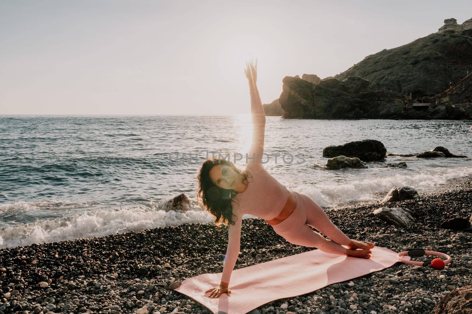 Woman sea yoga. Happy woman with black hair doing Pilates with the ring on the yoga mat near the sea on the pebble beach. Female fitness yoga concept. Healthy lifestyle, harmony and meditation. by panophotograph